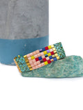 Peut inclure: Un bracelet de perles colorées avec un motif géométrique. Le bracelet présente un mélange de perles roses, jaunes, rouges, bleues et irisées, avec des accents dorés. Il est présenté sur une pierre vert clair.