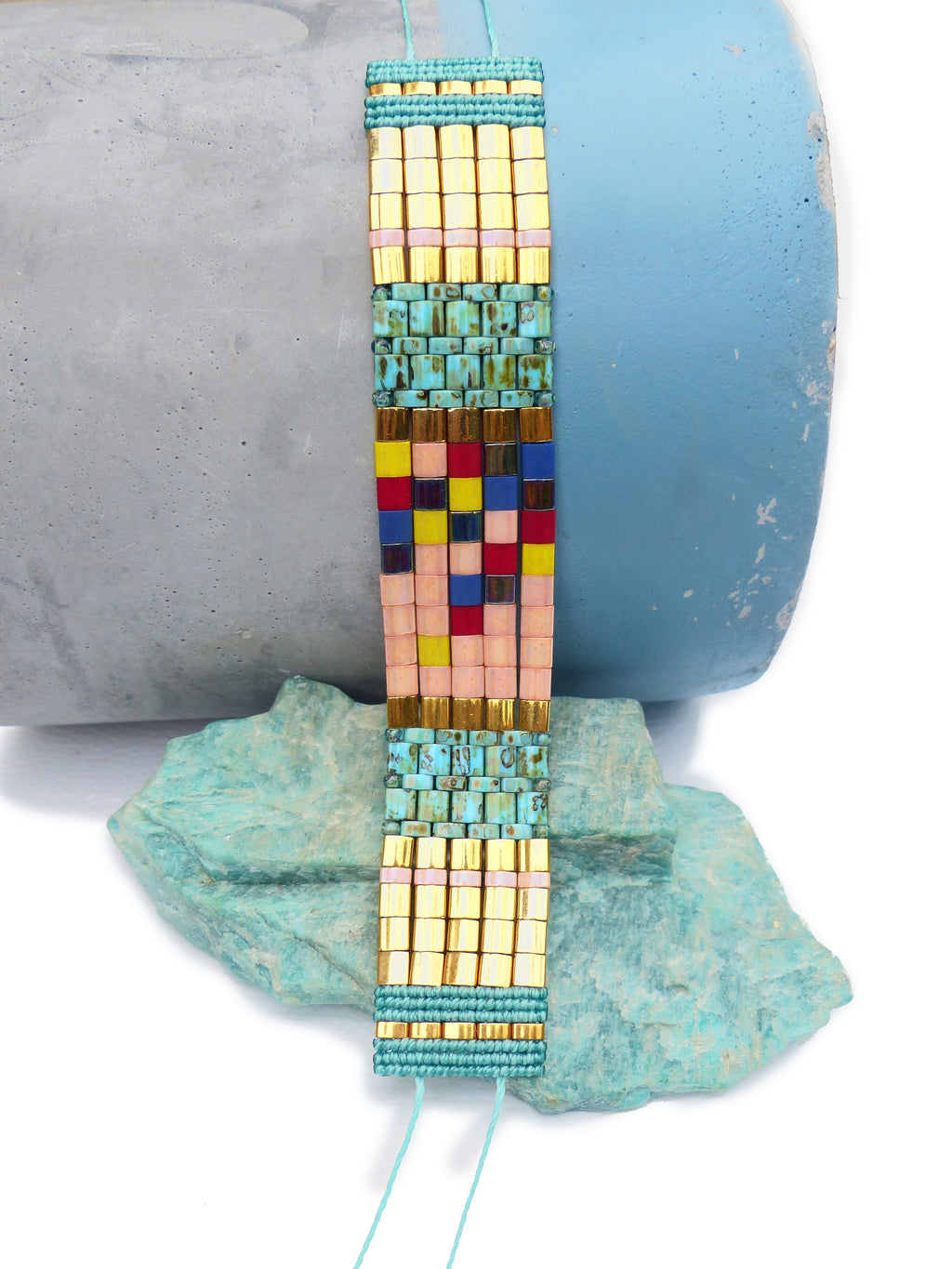 Peut inclure: Un bracelet de perles colorées avec des perles carrées dorées, turquoise, roses, rouges, jaunes et bleues. Le bracelet est tissé et possède une fermeture en cordon turquoise. Le bracelet mesure environ 2,5 cm de large.