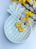 Peut inclure: Une paire de boucles d'oreilles en perles jaunes et dorées avec un design en frange, présentée sur un plat en céramique bleu en forme d'ananas.