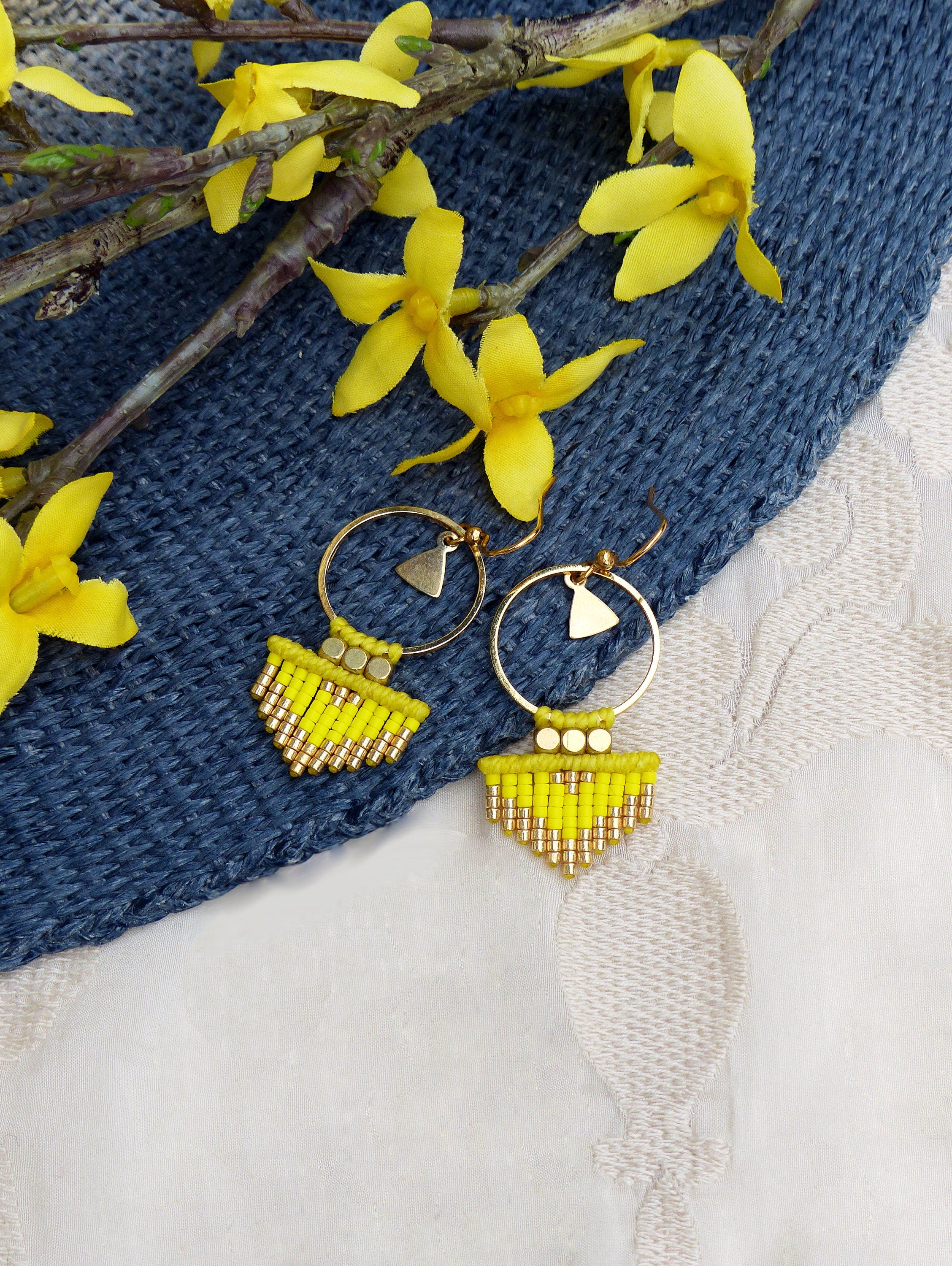 Peut inclure: Une paire de boucles d'oreilles créoles dorées avec des franges de perles jaunes. Les boucles d'oreilles sont sur une surface texturée bleue et blanche.
