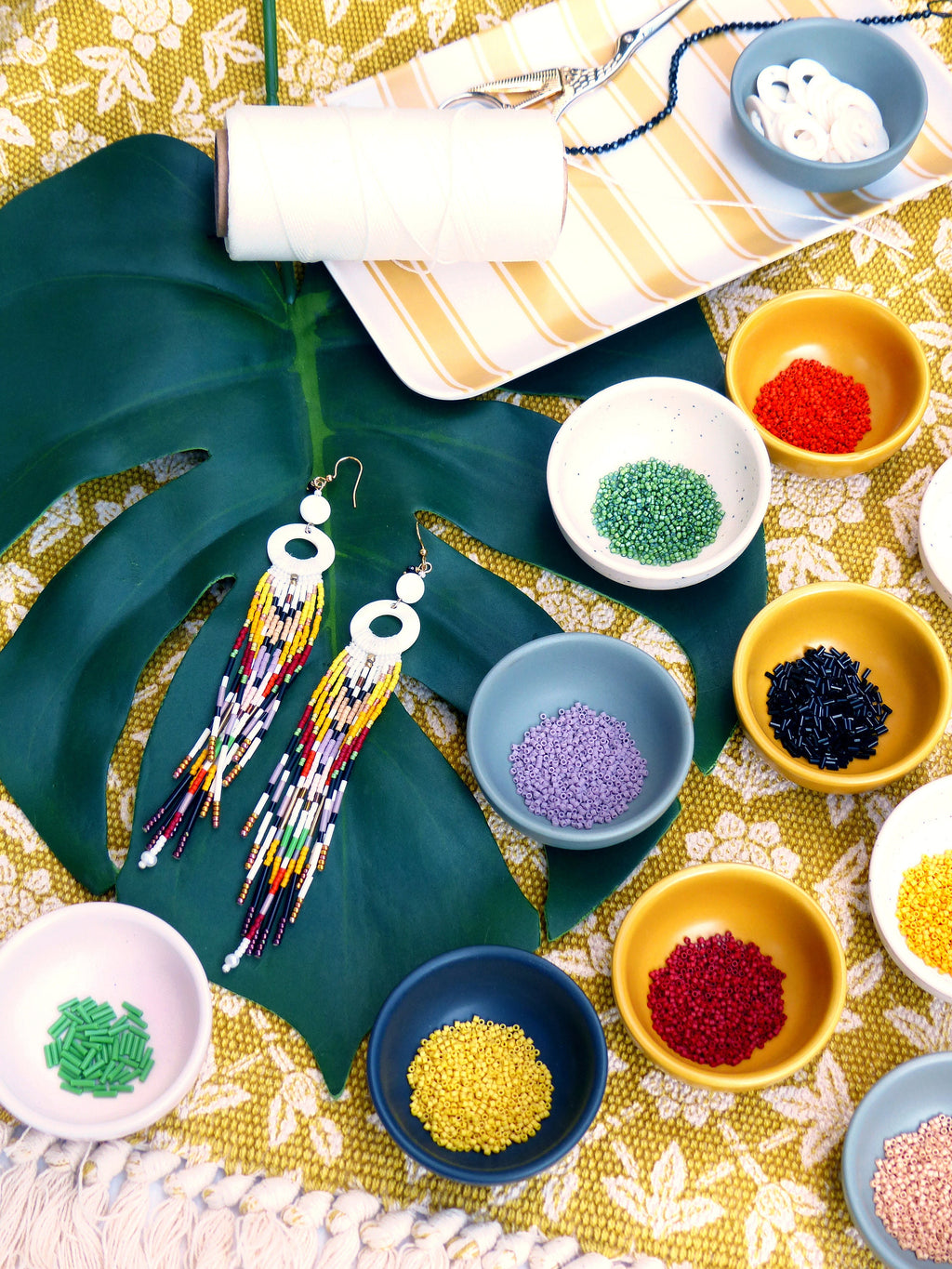 Peut inclure: Deux paires de boucles d'oreilles en perles colorées avec des accents circulaires blancs, disposées sur une feuille verte avec divers petits bols de perles colorées de différentes couleurs, notamment rouge, vert, noir, violet, jaune et orange.
