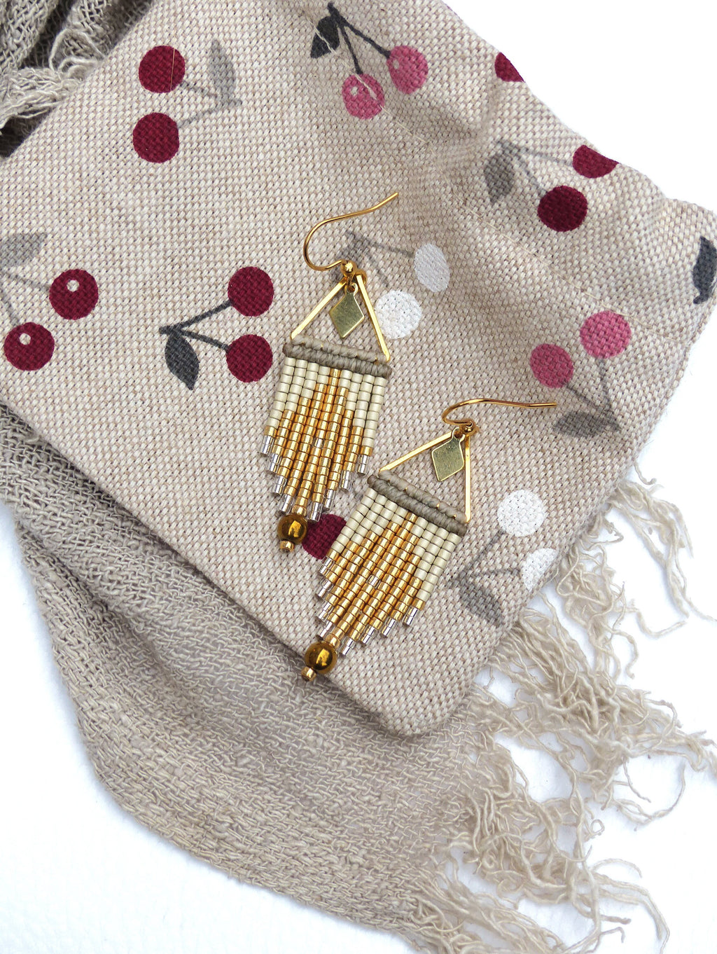 Peut inclure: Une paire de boucles d'oreilles en franges de perles dorées et crème avec un motif géométrique. Les boucles d'oreilles sont suspendues à un tissu beige à motif de cerises.