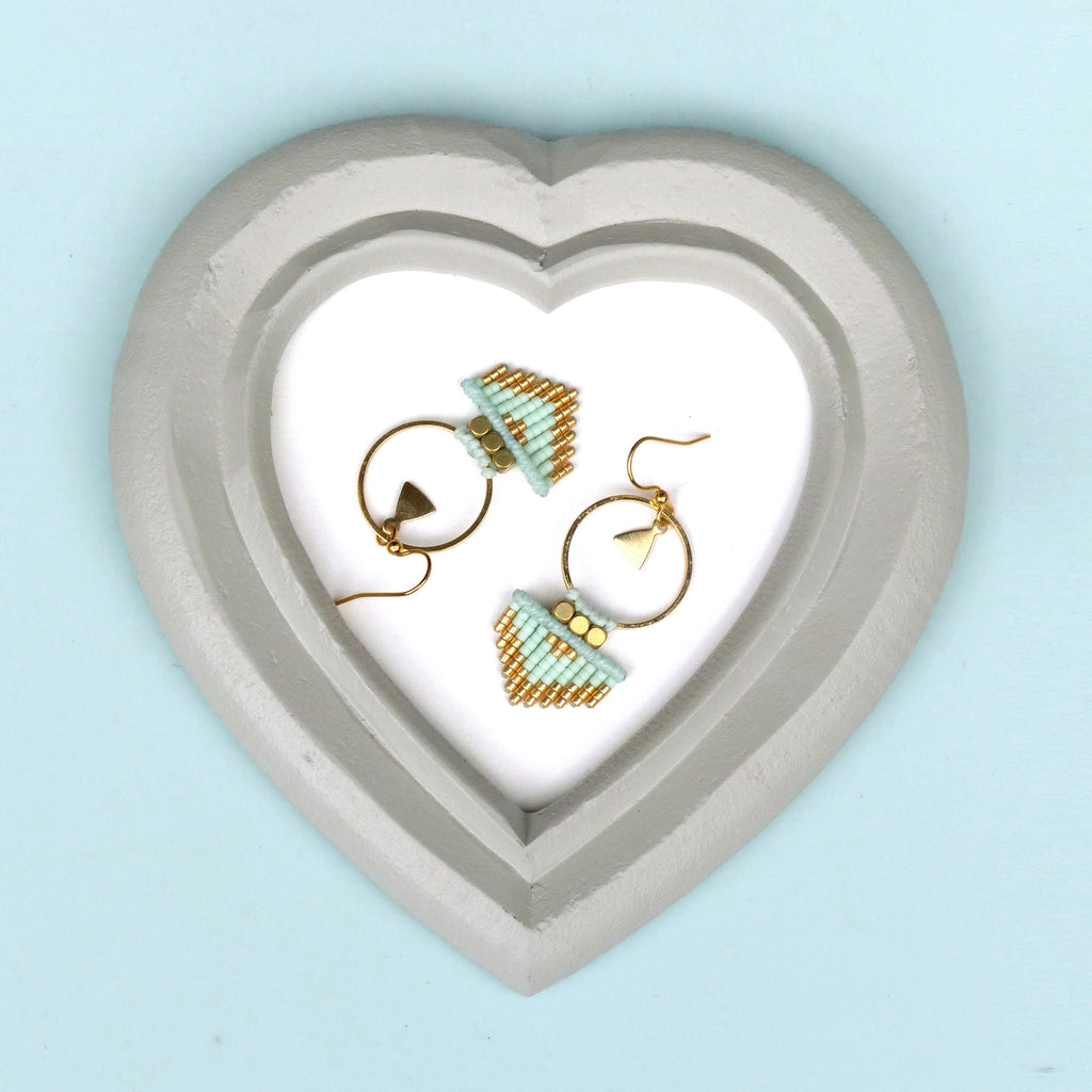 Peut inclure: Une paire de boucles d'oreilles en perles dorées et bleu clair avec un motif géométrique. Les boucles d'oreilles sont présentées dans un plat en forme de cœur gris.