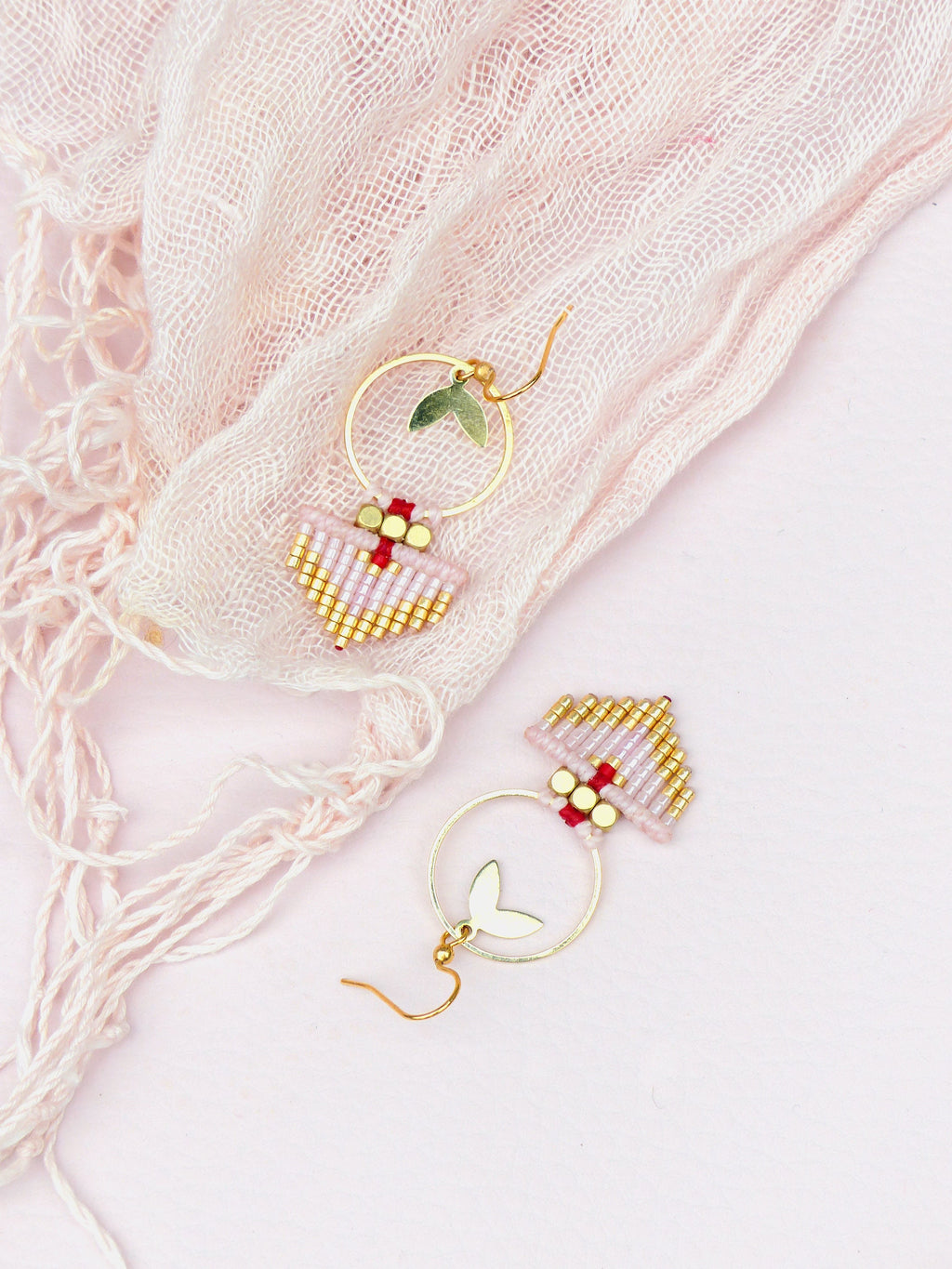 Peut inclure: Une paire de boucles d'oreilles créoles dorées avec un petit charme en forme de cœur et une frange de perles roses et dorées. Les boucles d'oreilles sont sur un fond rose.