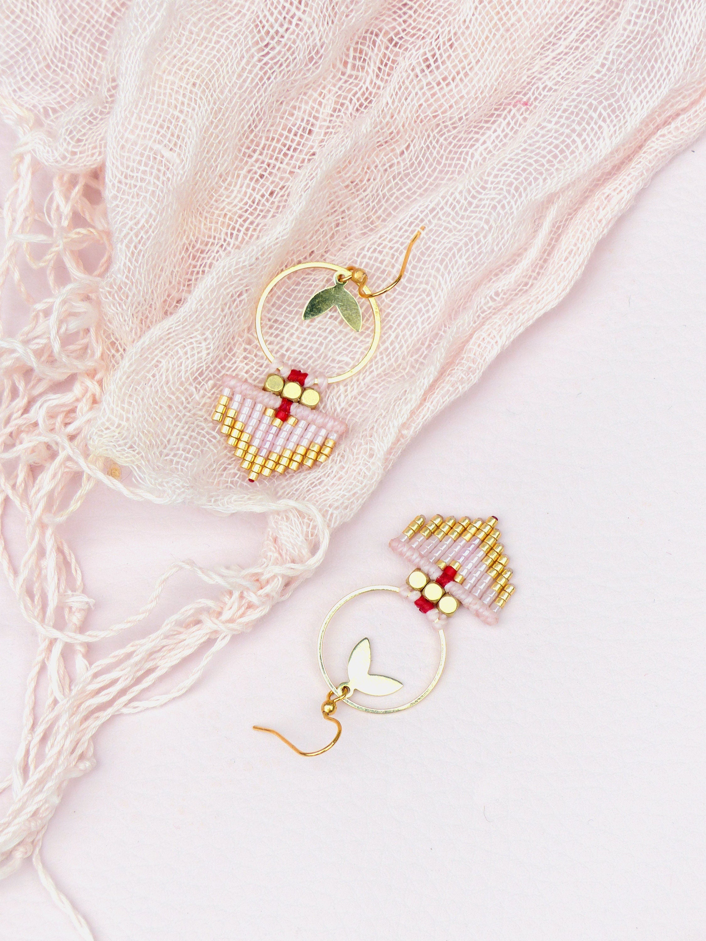 Peut inclure: Une paire de boucles d'oreilles créoles dorées avec un petit charme en forme de cœur et une frange de perles roses et dorées. Les boucles d'oreilles sont sur un fond rose.