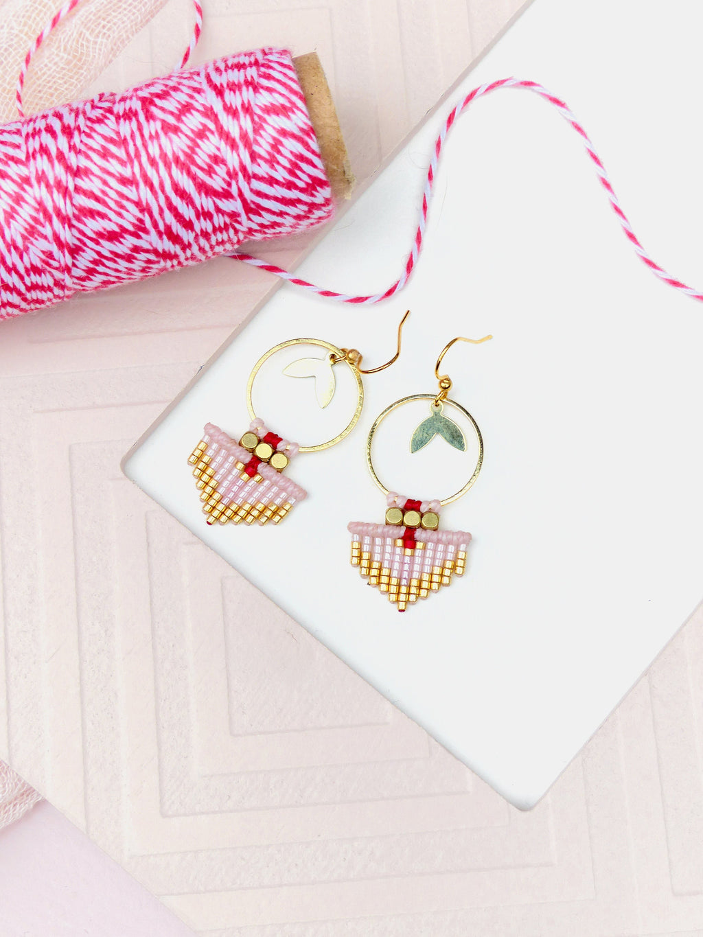 Peut inclure: Une paire de boucles d'oreilles créoles dorées avec des franges de perles roses et dorées et un petit charme de feuille verte. Les boucles d'oreilles sont sur une surface blanche avec une ficelle rouge et blanche.
