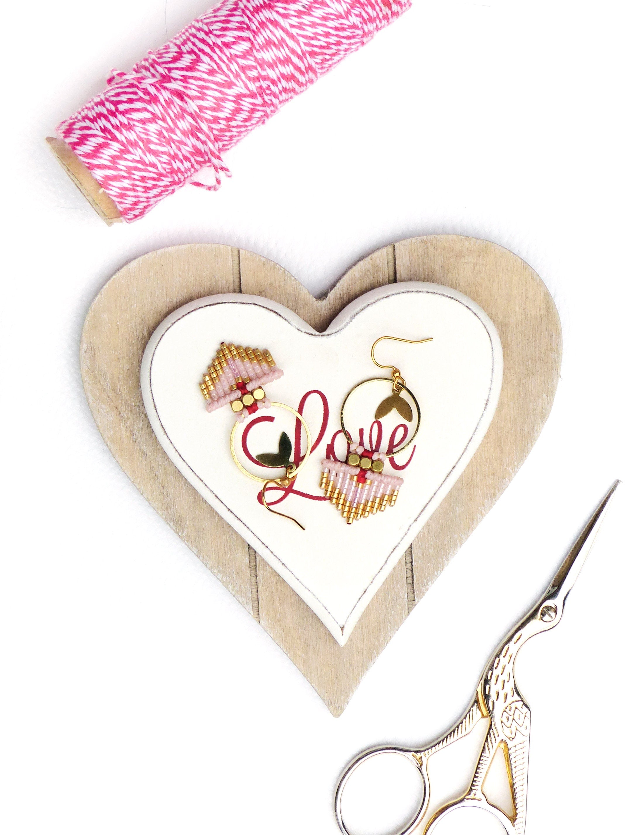 Peut inclure: Une paire de boucles d'oreilles en perles roses et dorées avec un charme en forme de cœur, exposées sur une plaque blanche en forme de cœur avec le mot "Love" écrit en rouge. La plaque est sur une base en bois en forme de cœur. Une paire de ciseaux dorés est également dans l'image.