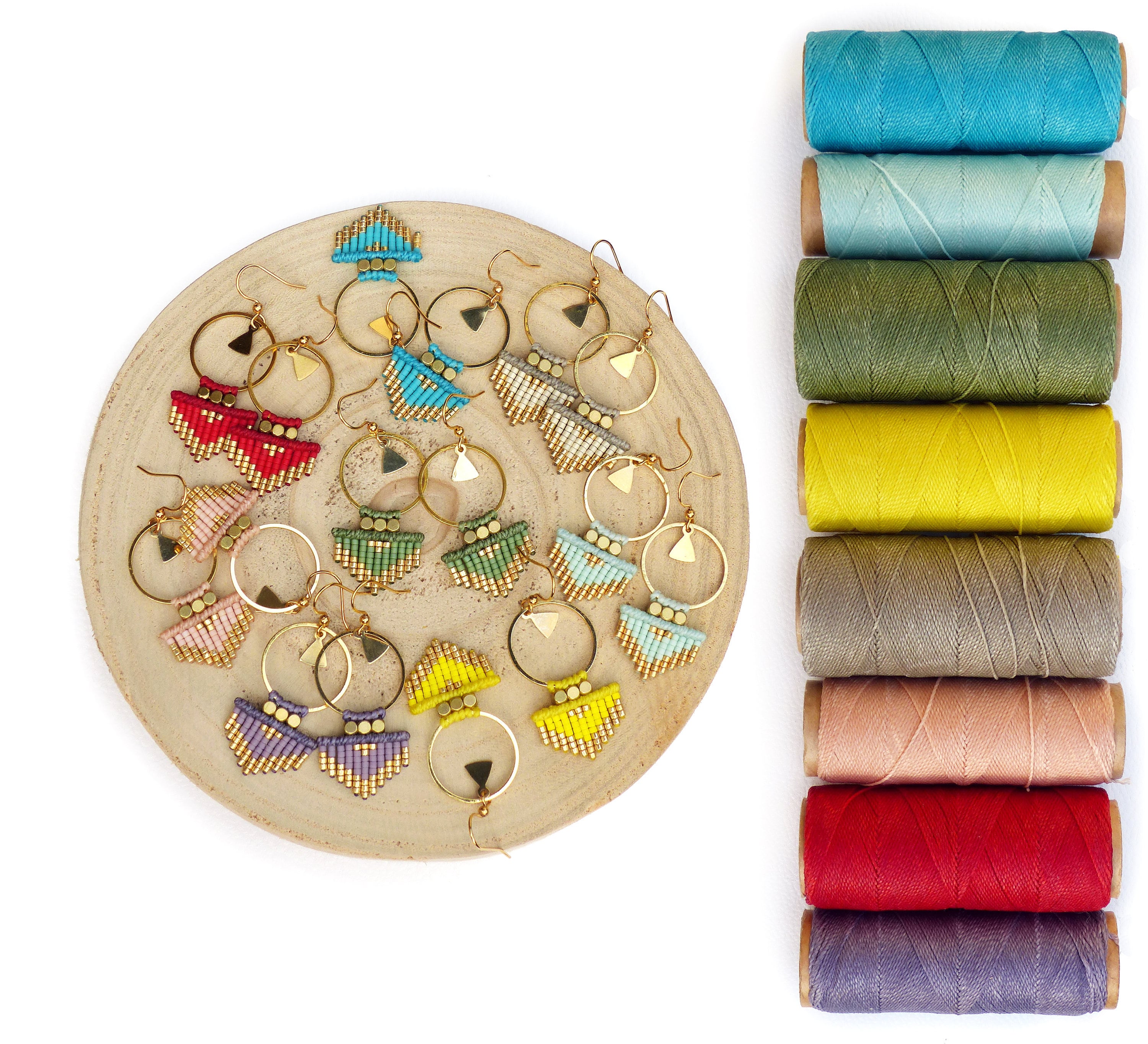 Peut inclure: Une collection de 12 paires de boucles d'oreilles faites à la main avec des motifs géométriques. Les boucles d'oreilles sont faites avec des perles colorées et des cercles dorés. Les boucles d'oreilles sont présentées sur une assiette en bois avec des bobines de fil de différentes couleurs.