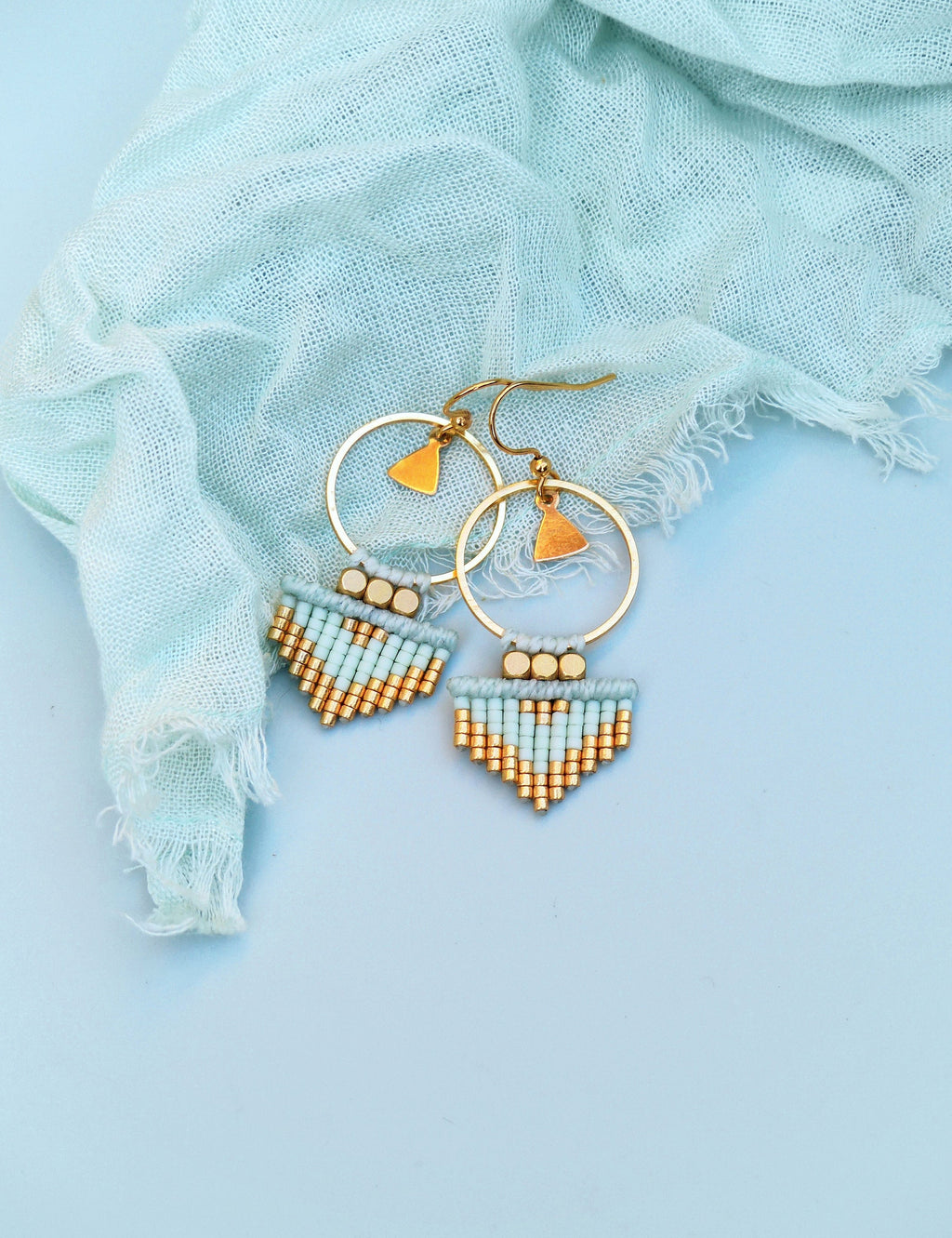 Peut inclure: Une paire de boucles d'oreilles créoles dorées avec une délicate frange de perles bleu clair et dorées. Les boucles d'oreilles sont sur un fond bleu clair.