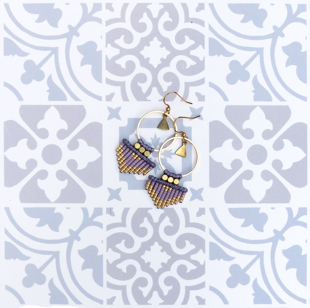 Peut inclure: Une paire de boucles d'oreilles en perles violettes et dorées avec des cercles dorés et des breloques triangulaires. Les boucles d'oreilles sont sur un fond blanc avec un motif géométrique.