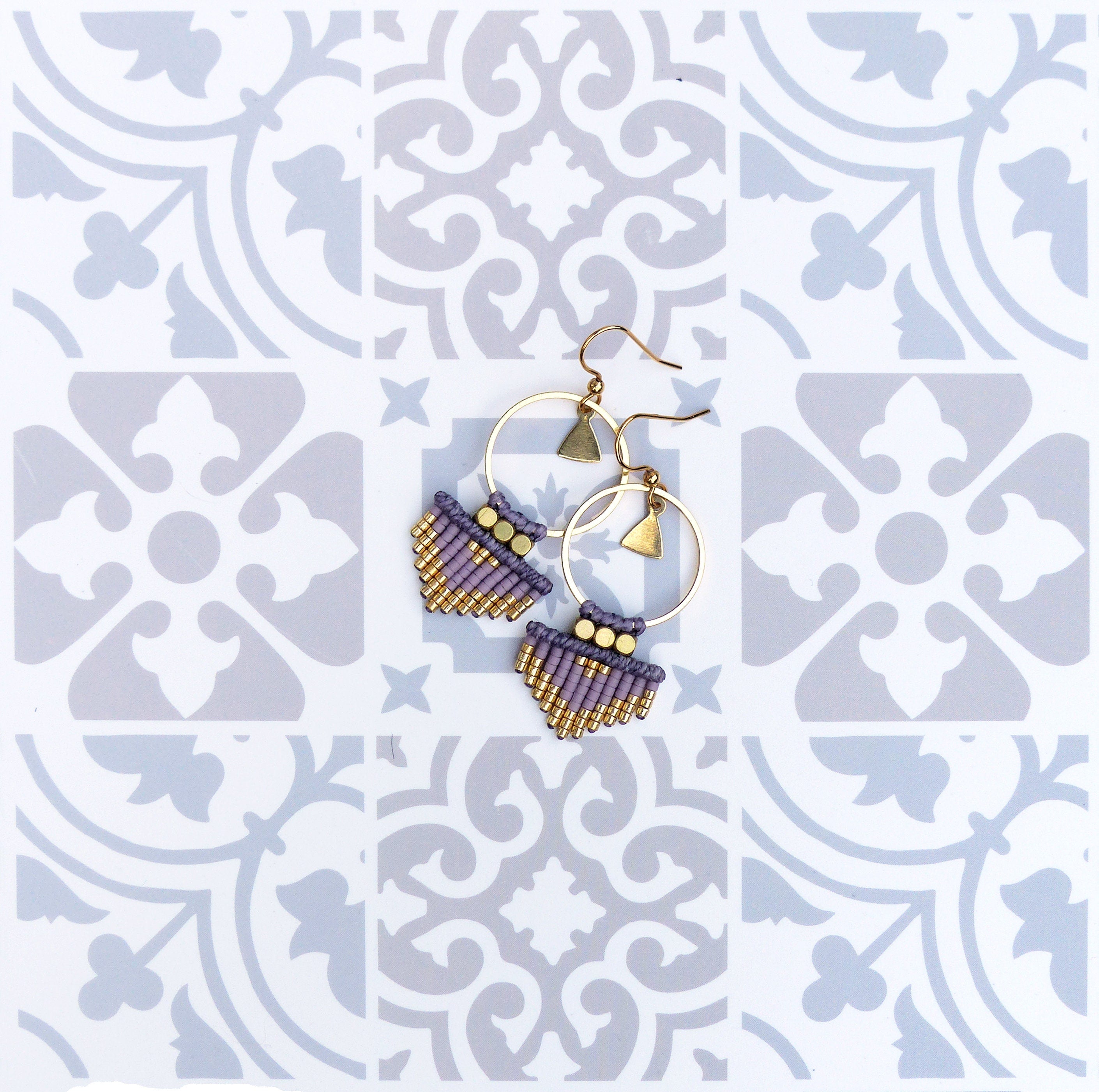 Peut inclure: Une paire de boucles d'oreilles en perles violettes et dorées avec des cercles dorés et des breloques triangulaires. Les boucles d'oreilles sont sur un fond blanc avec un motif géométrique.