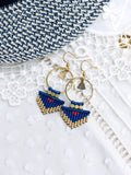 Peut inclure: Une paire de boucles d'oreilles créoles dorées avec des franges de perles bleues et dorées. Les boucles d'oreilles ont un petit charme triangulaire suspendu à la créole.