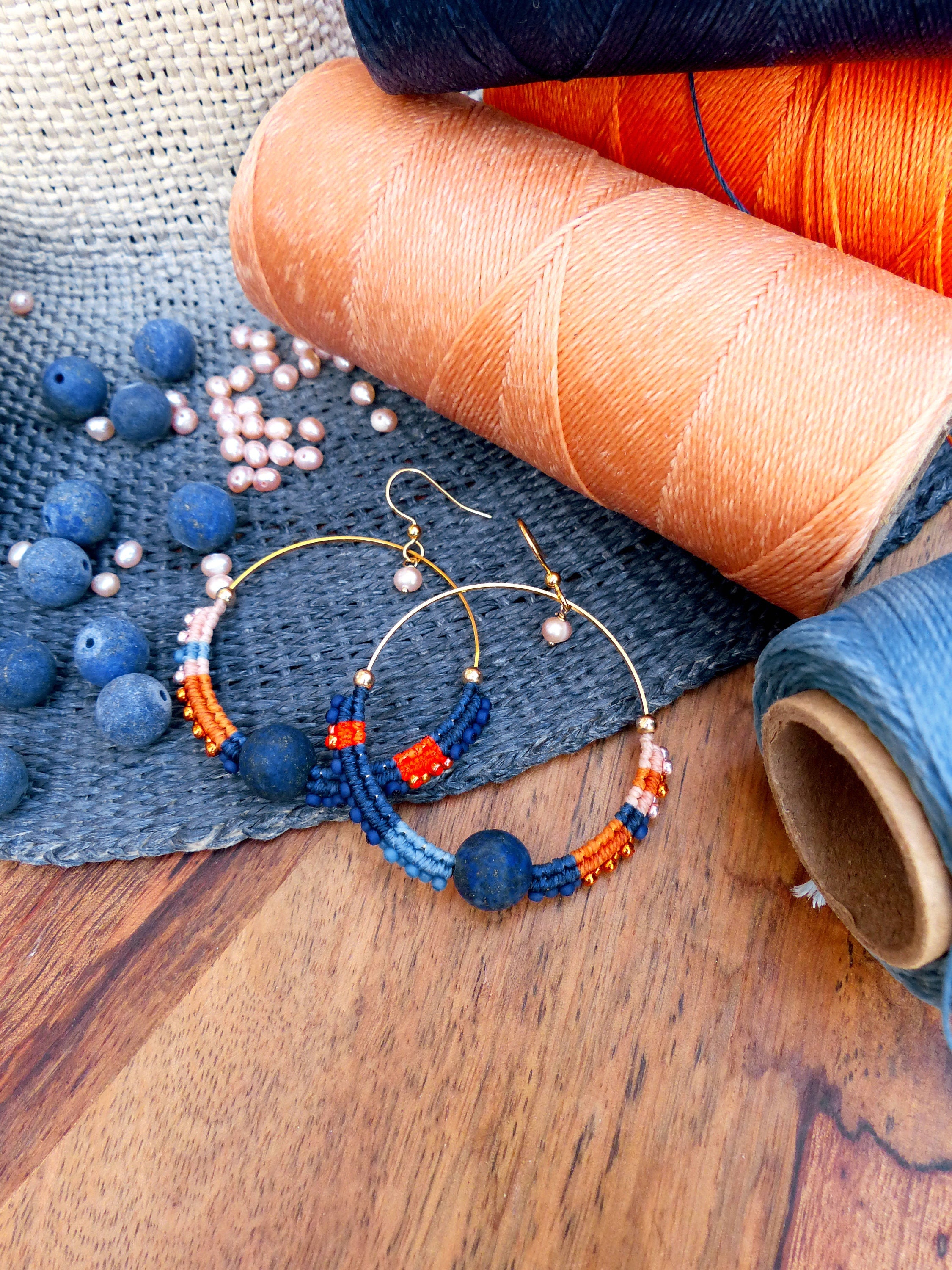 Peut inclure: Une paire de boucles d'oreilles créoles faites à la main avec un design en macramé bleu, orange et blanc. Chaque boucle d'oreille est ornée d'une grosse perle de pierre bleu foncé.