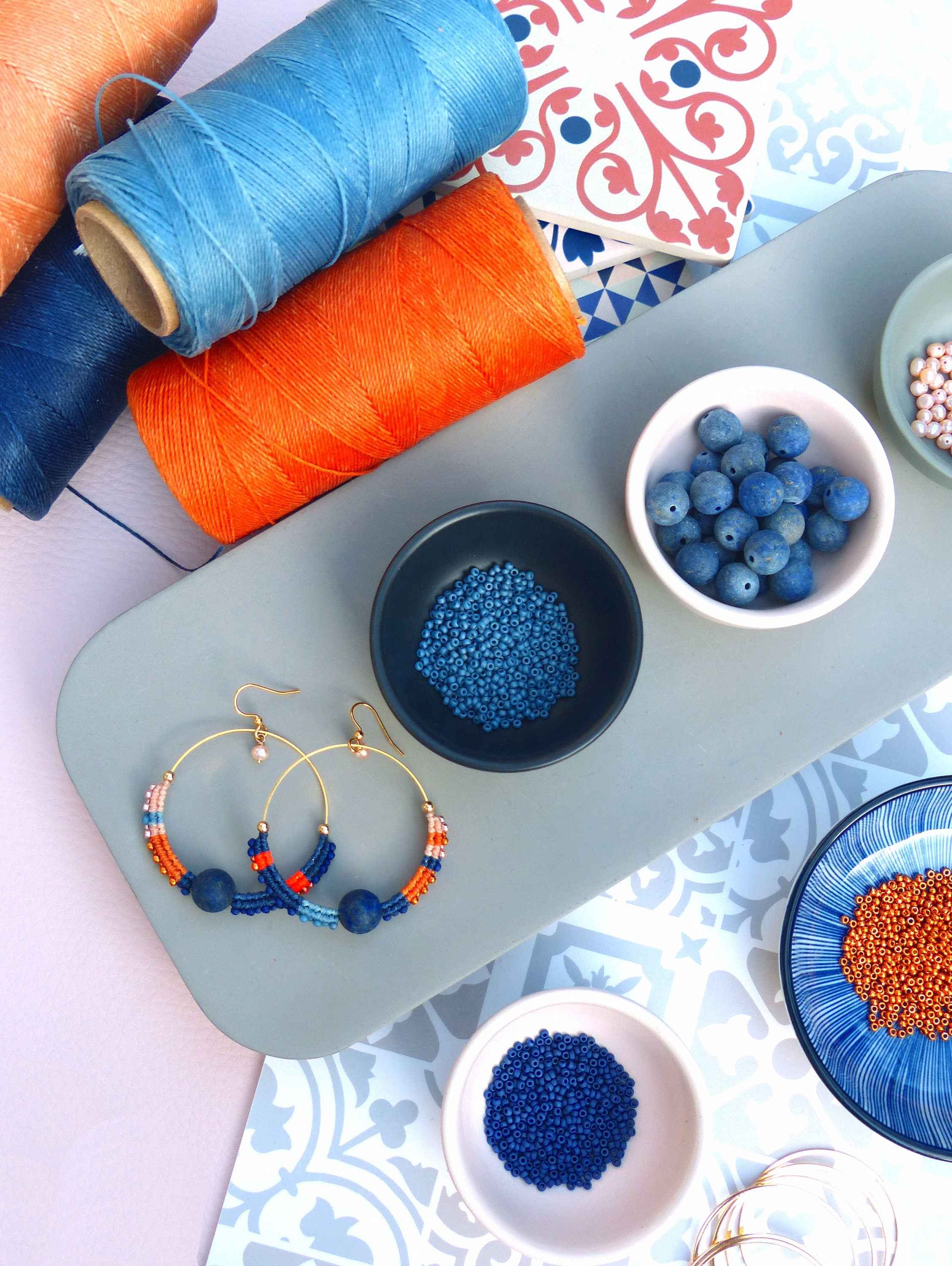 Peut inclure: Un plateau avec du fil à broder bleu et orange, des perles bleues et orange, et une paire de boucles d'oreilles créoles avec des perles bleues, orange et roses. Les boucles d'oreilles sont sur une surface grise avec un fond à motifs bleus et blancs.