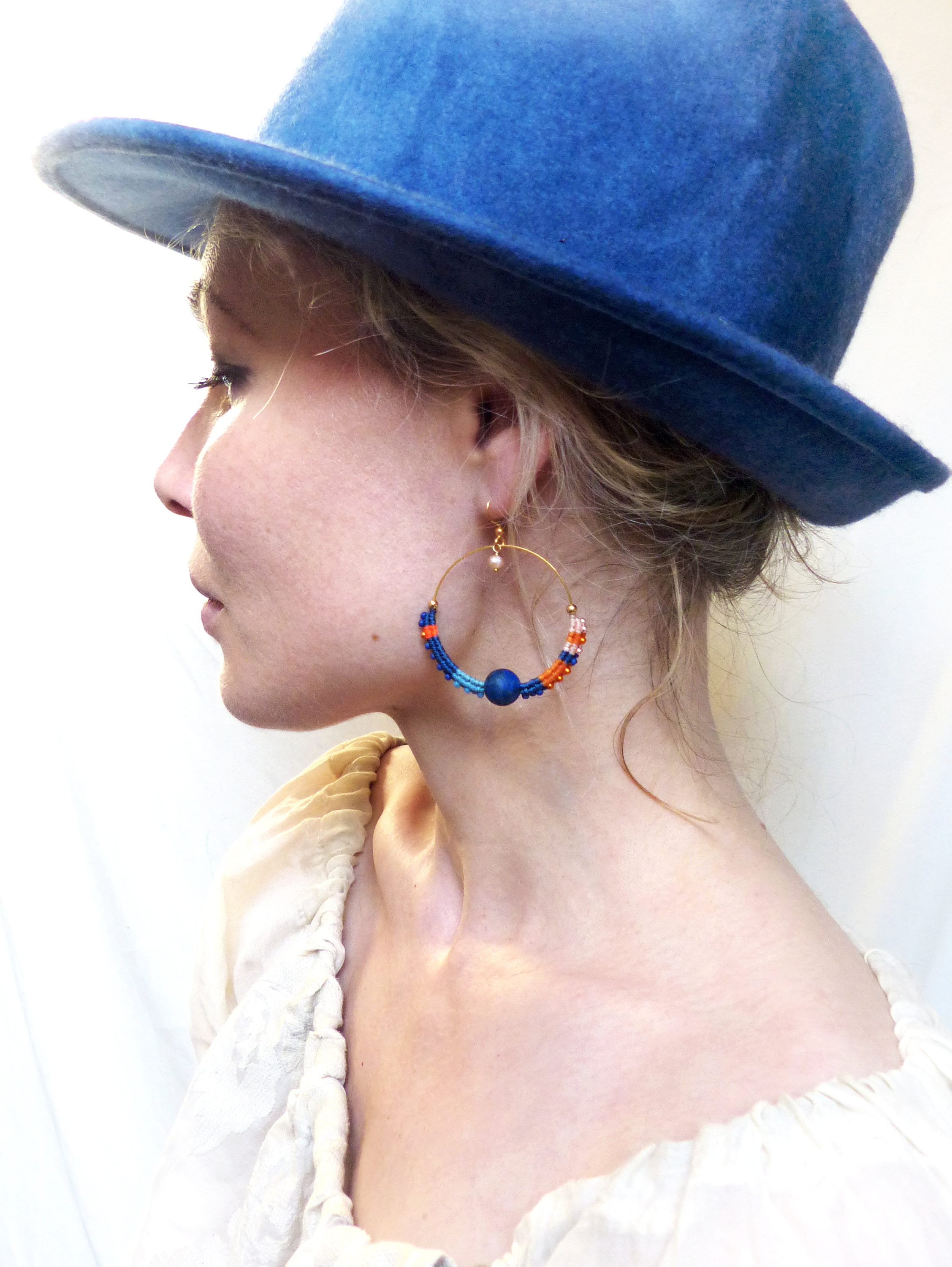Peut inclure: Une femme portant un chapeau bleu et une blouse blanche avec une paire de boucles d'oreilles créoles en perles bleues, orange et blanches.