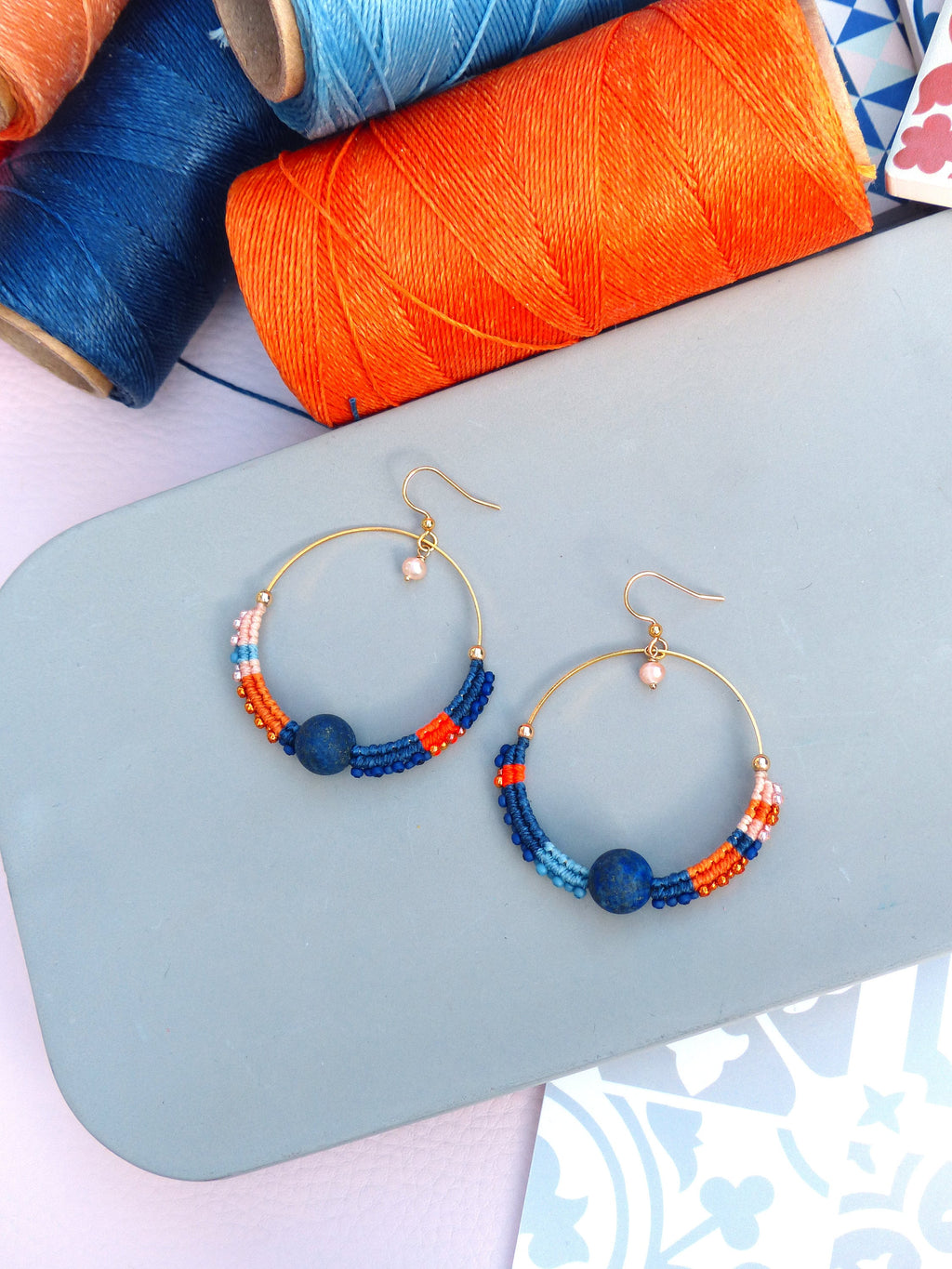 Peut inclure: Une paire de boucles d'oreilles créoles dorées avec des perles bleues, orange et roses. Les boucles d'oreilles sont sur une surface bleu clair.