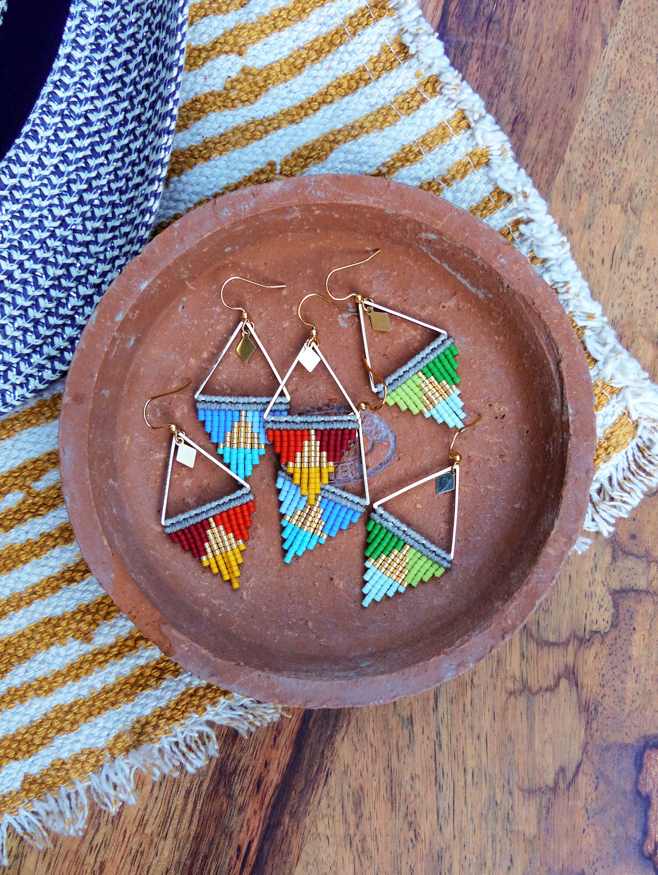 Peut inclure: Quatre paires de boucles d'oreilles triangulaires en perles colorées avec des accents dorés. Les boucles d'oreilles sont présentées sur une assiette en terre cuite.