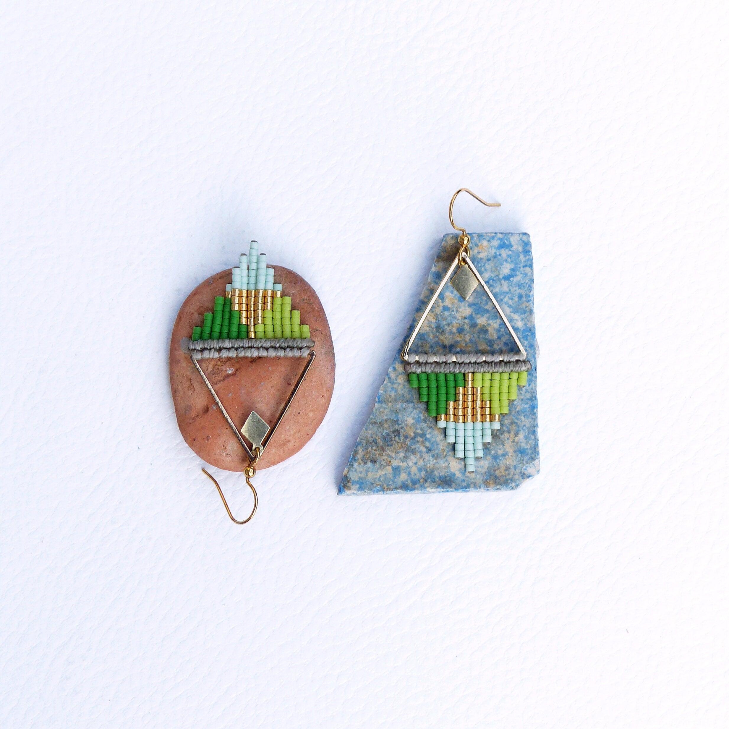 Peut inclure: Une paire de boucles d'oreilles géométriques de forme triangulaire. Les boucles d'oreilles sont faites de perles vertes, bleues et dorées et ont un cadre en fil d'argent. Les boucles d'oreilles sont présentées sur une pierre bleue et brune.