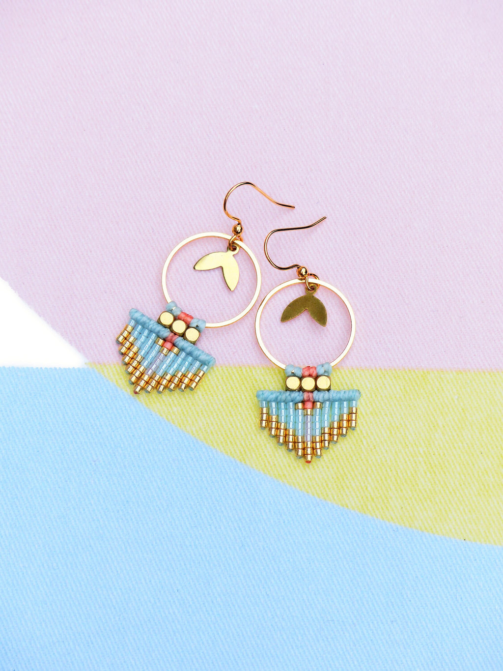Peut inclure: Boucles d'oreilles dorées avec un charme en forme de feuille et un design à franges de perles turquoise et dorées. Les boucles d'oreilles ont une section supérieure perlée rose et dorée. Le fond est un bloc de couleurs pastel rose, jaune et bleu.