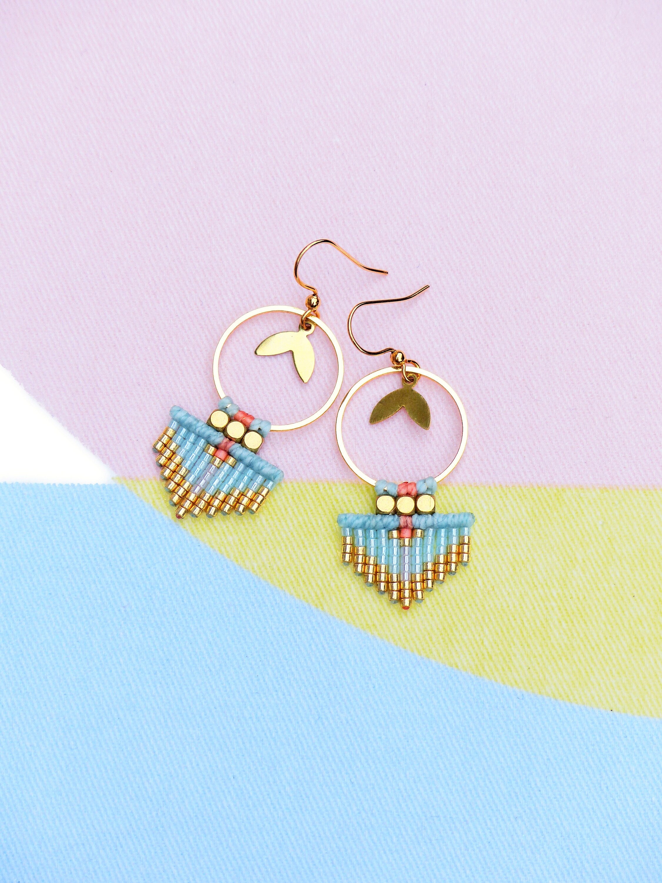 Peut inclure: Boucles d'oreilles dorées avec un charme en forme de feuille et un design à franges de perles turquoise et dorées. Les boucles d'oreilles ont une section supérieure perlée rose et dorée. Le fond est un bloc de couleurs pastel rose, jaune et bleu.