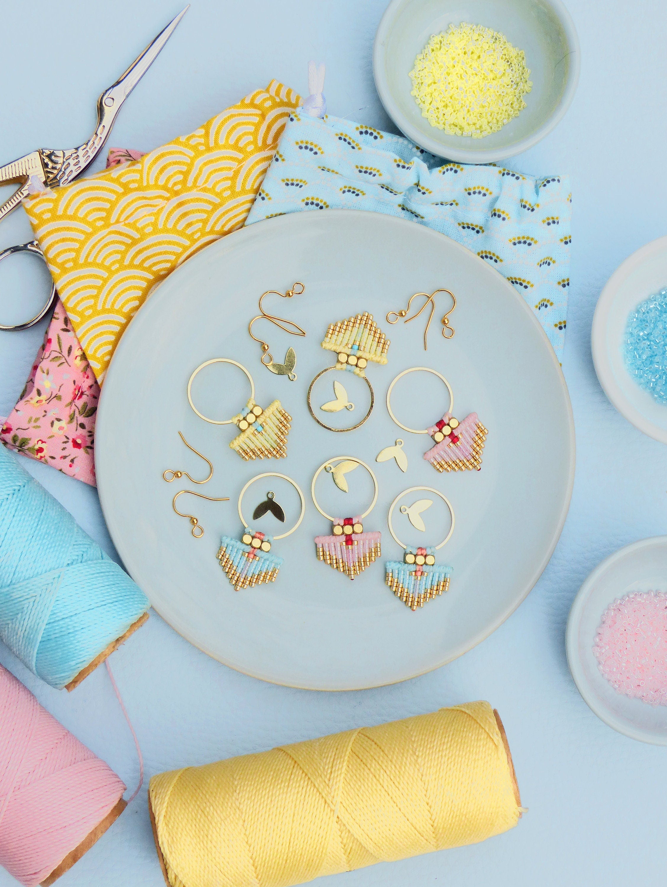 Peut inclure: Boucles d'oreilles faites à la main avec des cerceaux dorés et des motifs perlés aux couleurs pastel, disposées sur une assiette bleu clair. Les boucles d'oreilles présentent des accents dorés et des breloques en forme d'oiseau. Des bobines de fil colorées et des pochettes en tissu entourent l'assiette.