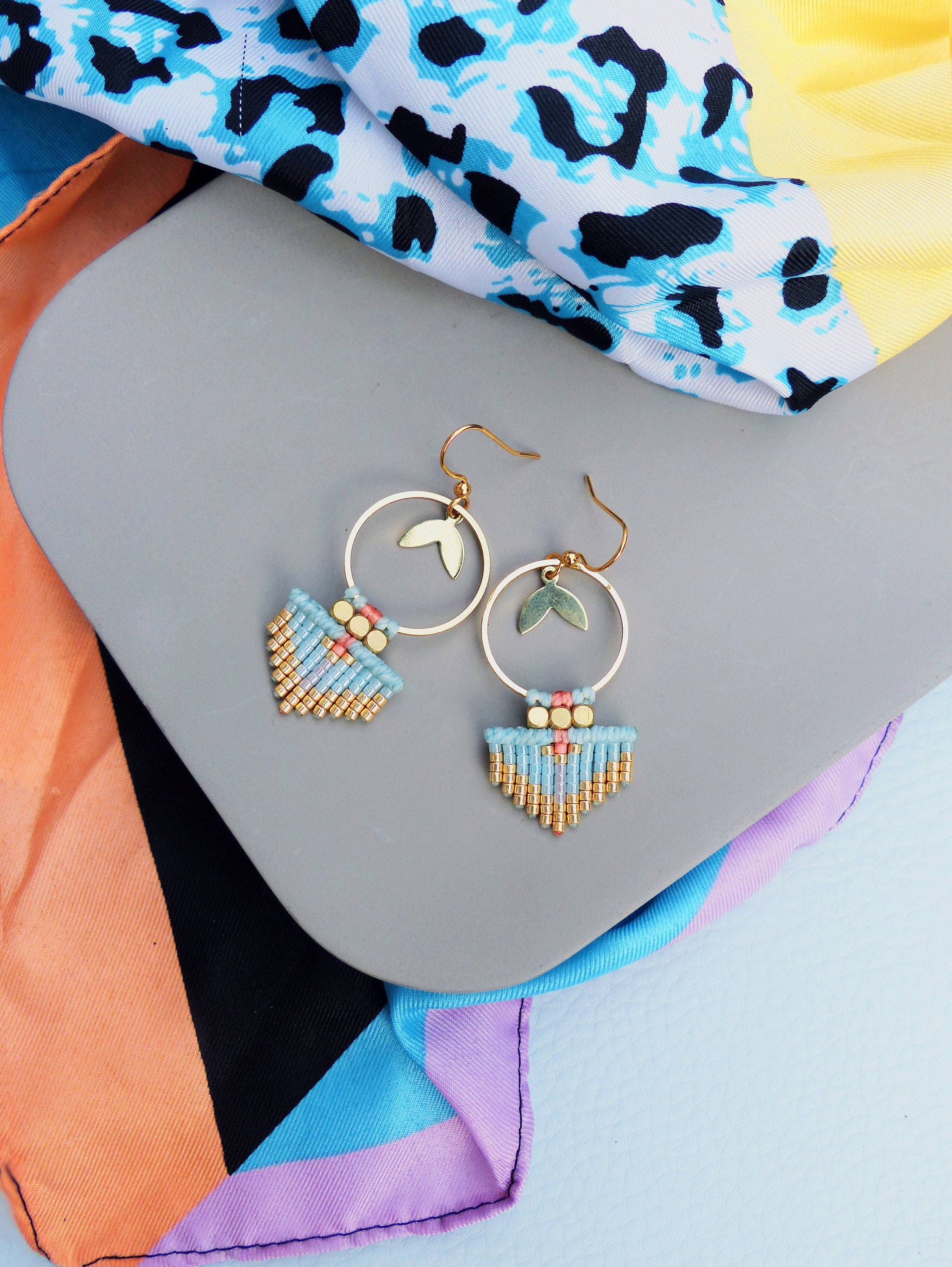 Peut inclure: Boucles d'oreilles dorées avec un design circulaire et des accents de feuilles. Les boucles d'oreilles présentent une frange de perles bleu clair et dorées. Un foulard à motifs colorés est en arrière-plan.