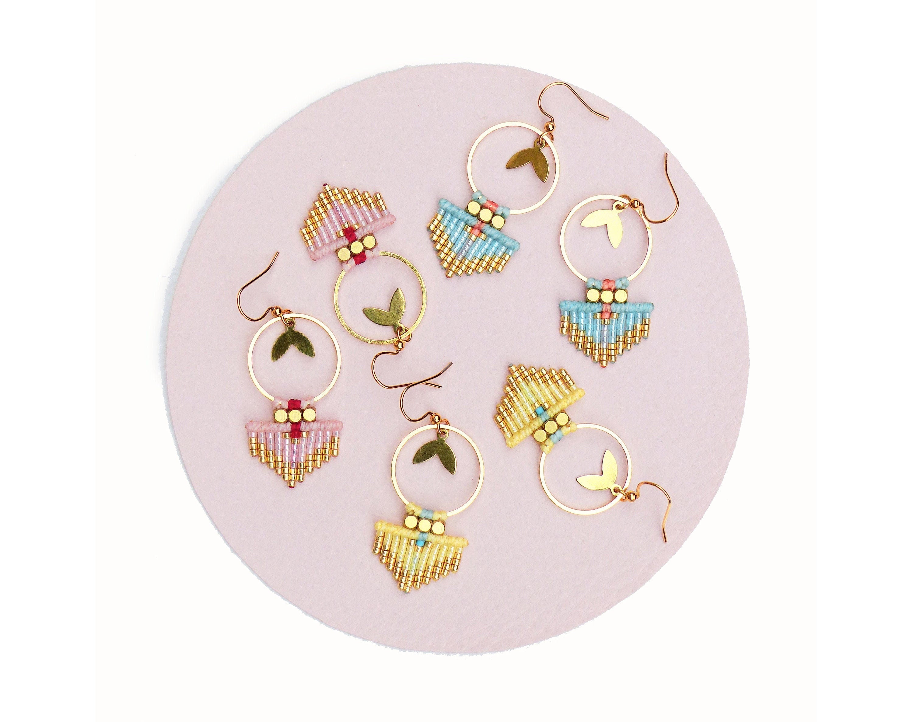 Peut inclure: Plusieurs boucles d'oreilles faites à la main de style bohème. Chaque boucle d'oreille est composée d'un cercle doré, d'un charm en forme de feuille et d'un motif de perles roses, bleues et jaunes. Les boucles d'oreilles sont présentées sur une surface rose.