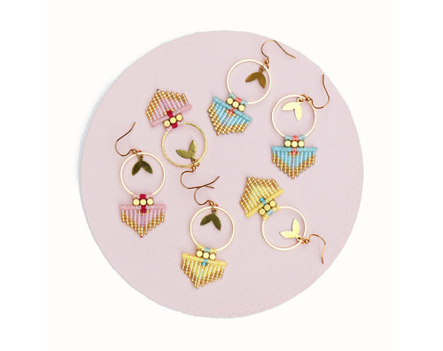 Peut inclure: Plusieurs boucles d'oreilles faites à la main de style bohème. Chaque boucle d'oreille est composée d'un cercle doré, d'un charm en forme de feuille et d'un motif de perles roses, bleues et jaunes. Les boucles d'oreilles sont présentées sur une surface rose.