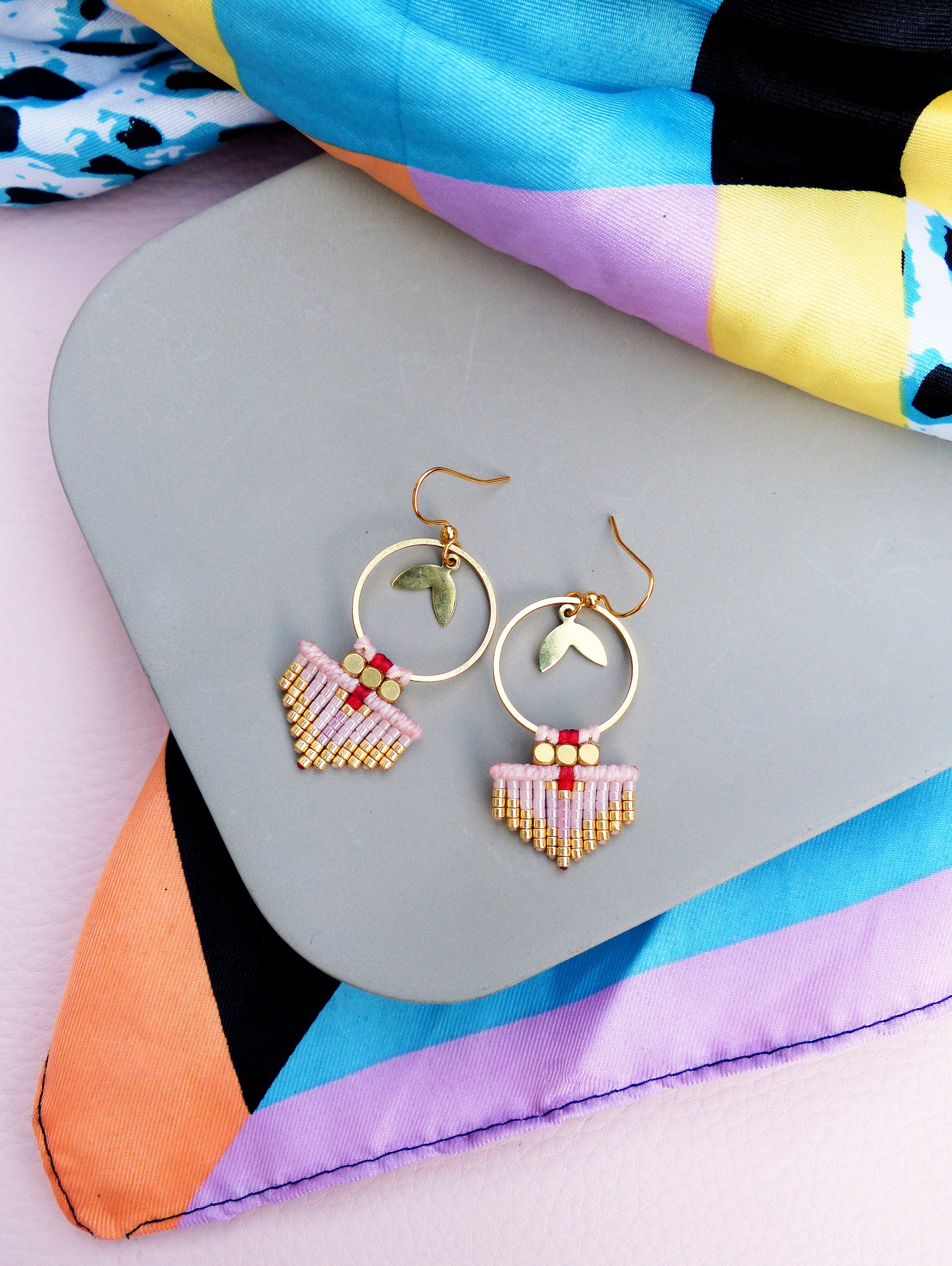 Peut inclure: Boucles d'oreilles créoles dorées avec un charme en forme de feuille et des franges perlées roses, dorées et rouges. Les boucles d'oreilles sont présentées sur une surface grise avec une écharpe colorée à motifs en arrière-plan. Style bohème.
