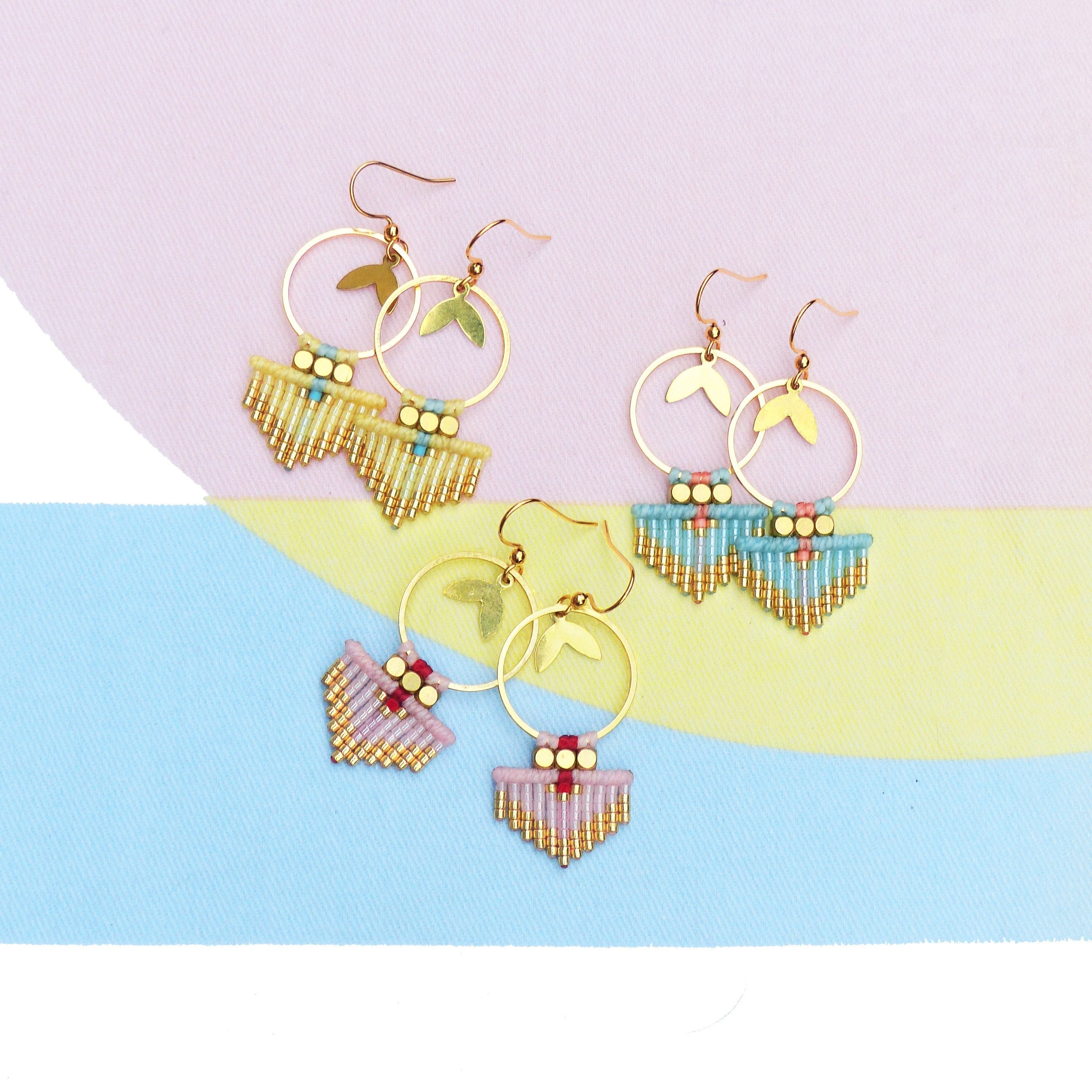 Peut inclure: Boucles d'oreilles dorées avec des accents de feuilles et des franges de perles jaunes, bleues et roses. Les boucles d'oreilles ont un design géométrique avec des perles dorées, bleues et rouges. Le fond est un bloc de couleurs pastel rose, jaune et bleu.
