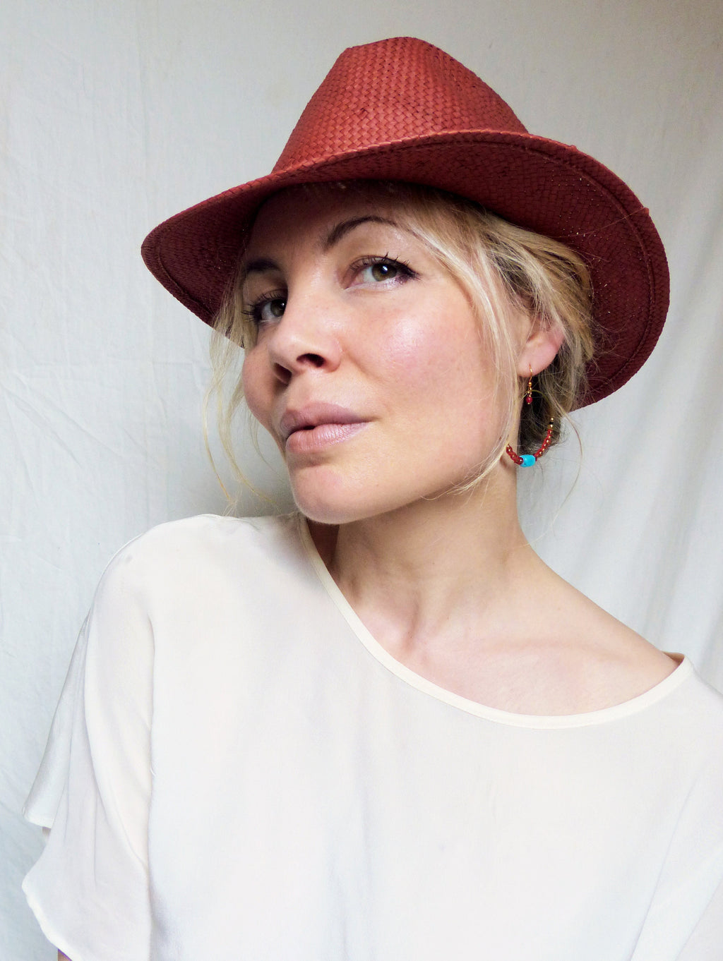 Peut inclure: Une femme portant une chemise blanche et un chapeau fedora en paille rouge. Elle porte des boucles d'oreilles créoles avec des perles turquoise et rouges.