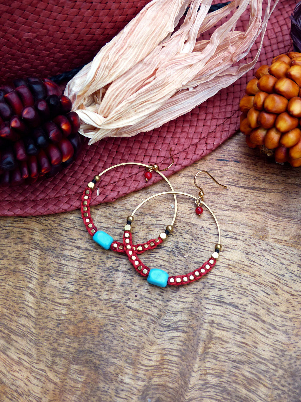 Peut inclure: Une paire de boucles d'oreilles créoles avec des perles rouges et dorées et des pierres turquoise. Les boucles d'oreilles sont sur une surface en bois.