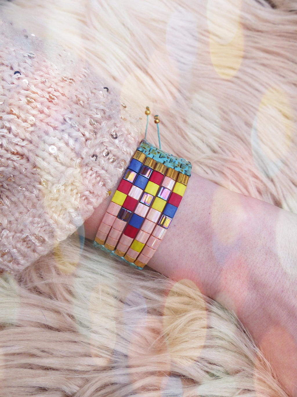 Peut inclure: Un bracelet de perles colorées avec des perles rectangulaires roses, bleues, jaunes et rouges. Le bracelet est porté sur un poignet, sur un fond doux et duveteux. Le bracelet a des accents dorés.