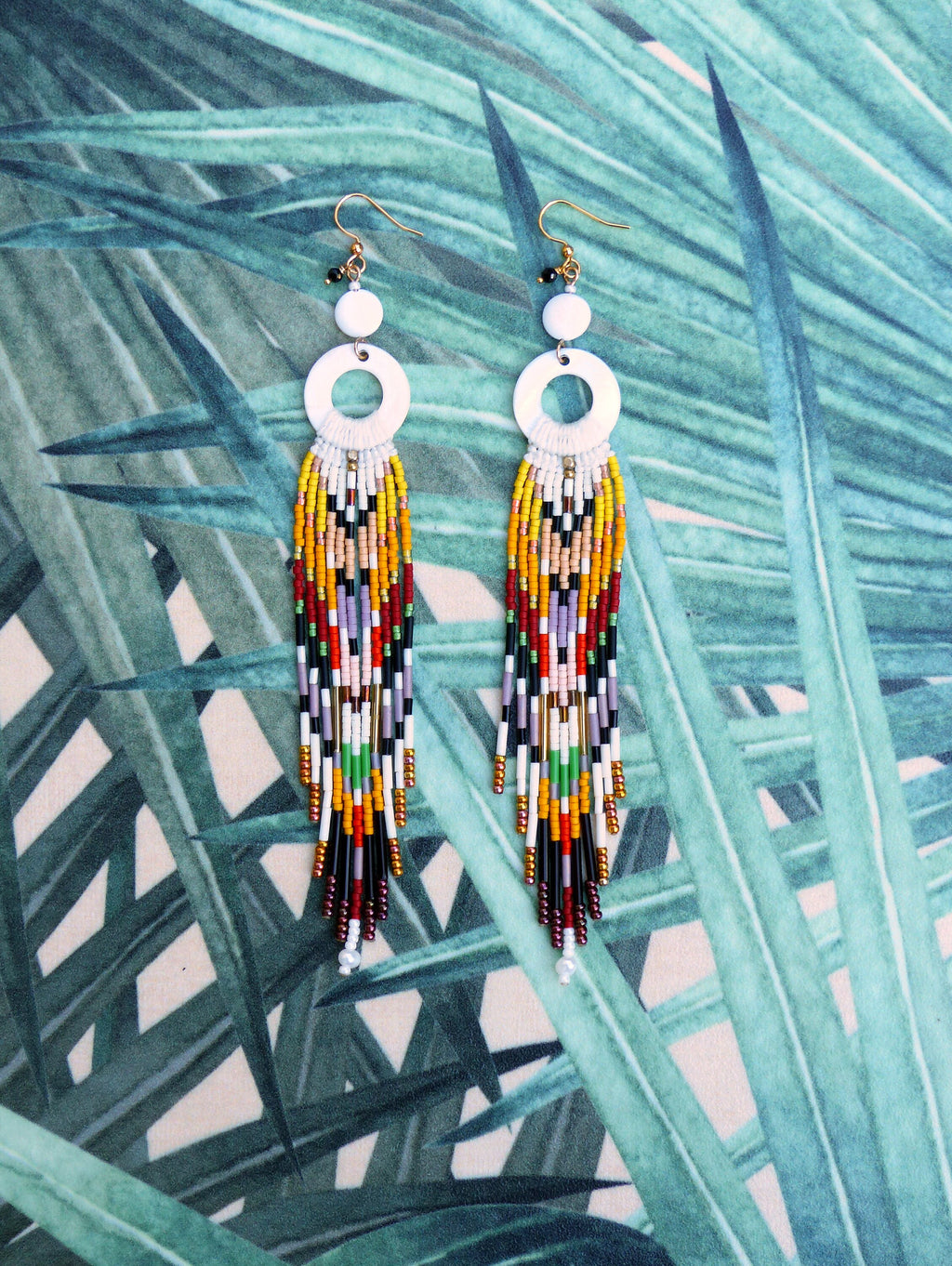 Peut inclure: Une paire de longues boucles d'oreilles en perles colorées avec des sommets circulaires blancs et des crochets dorés. Les boucles d'oreilles présentent une variété de couleurs, notamment le rouge, le jaune, le vert, le noir et le blanc.