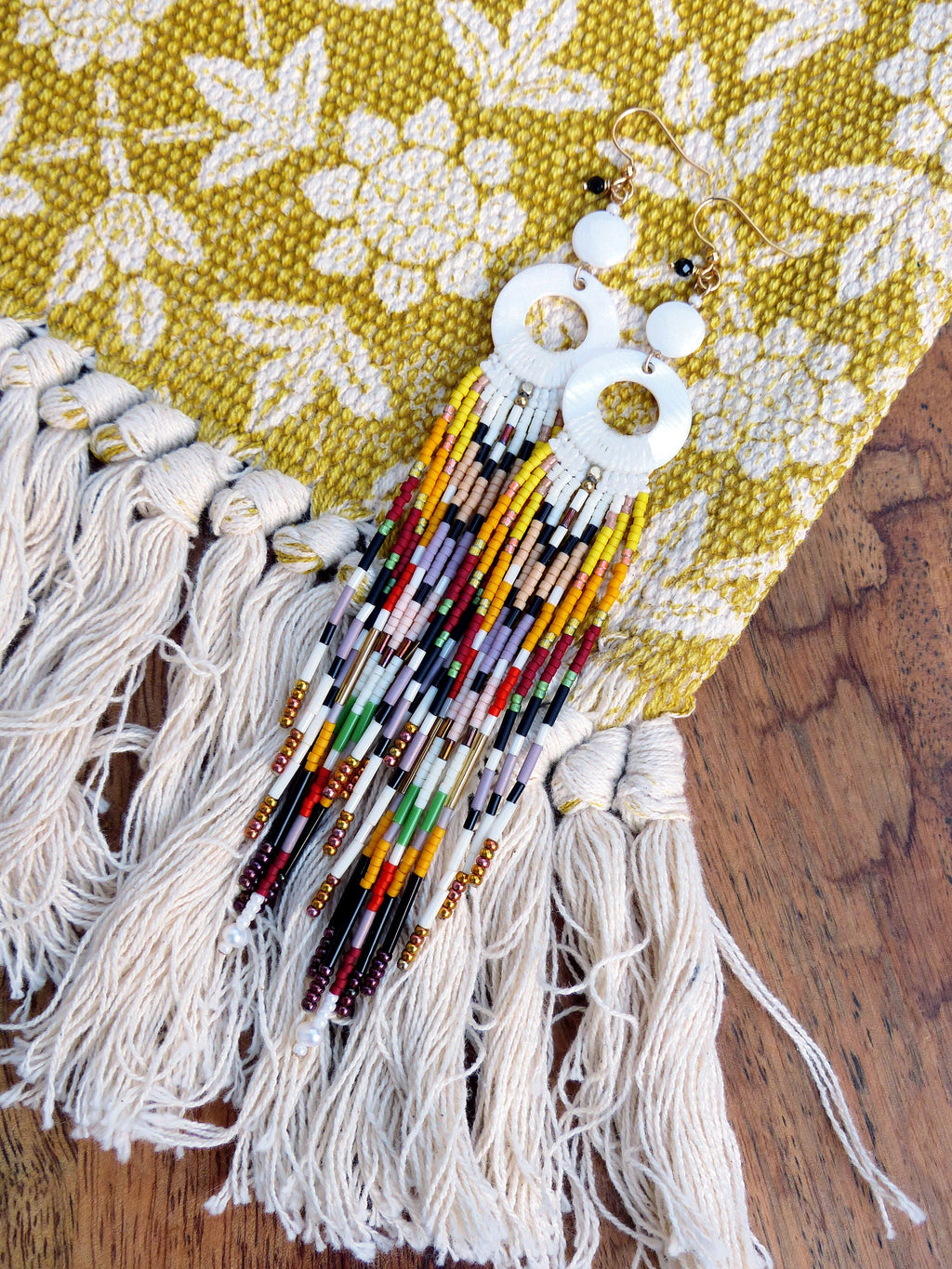 Peut inclure: Une paire de boucles d'oreilles rondes blanches avec une longue frange de perles. La frange est composée de perles colorées dans des tons de rouge, orange, jaune, vert, bleu, violet et noir. Les boucles d'oreilles sont suspendues à un fond en tissu blanc.