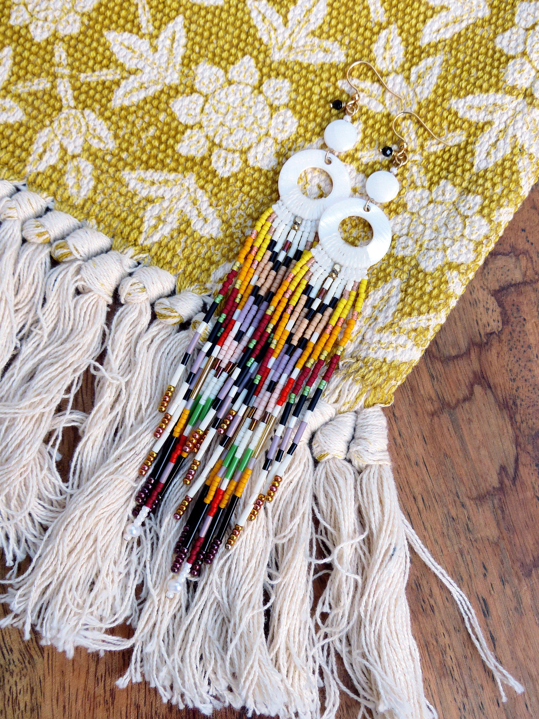 Peut inclure: Une paire de boucles d'oreilles rondes blanches avec une longue frange de perles. La frange est composée de perles colorées dans des tons de rouge, orange, jaune, vert, bleu, violet et noir. Les boucles d'oreilles sont suspendues à un fond en tissu blanc.