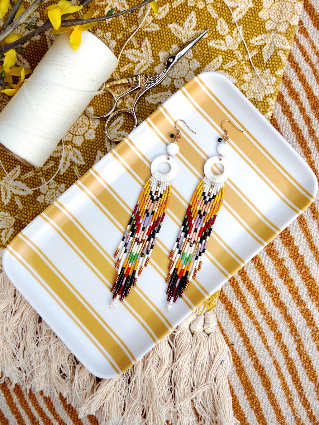 Peut inclure: Une paire de longues boucles d'oreilles en perles colorées avec des accents circulaires blancs. Les boucles d'oreilles sont présentées sur un plateau à rayures blanches et jaunes.