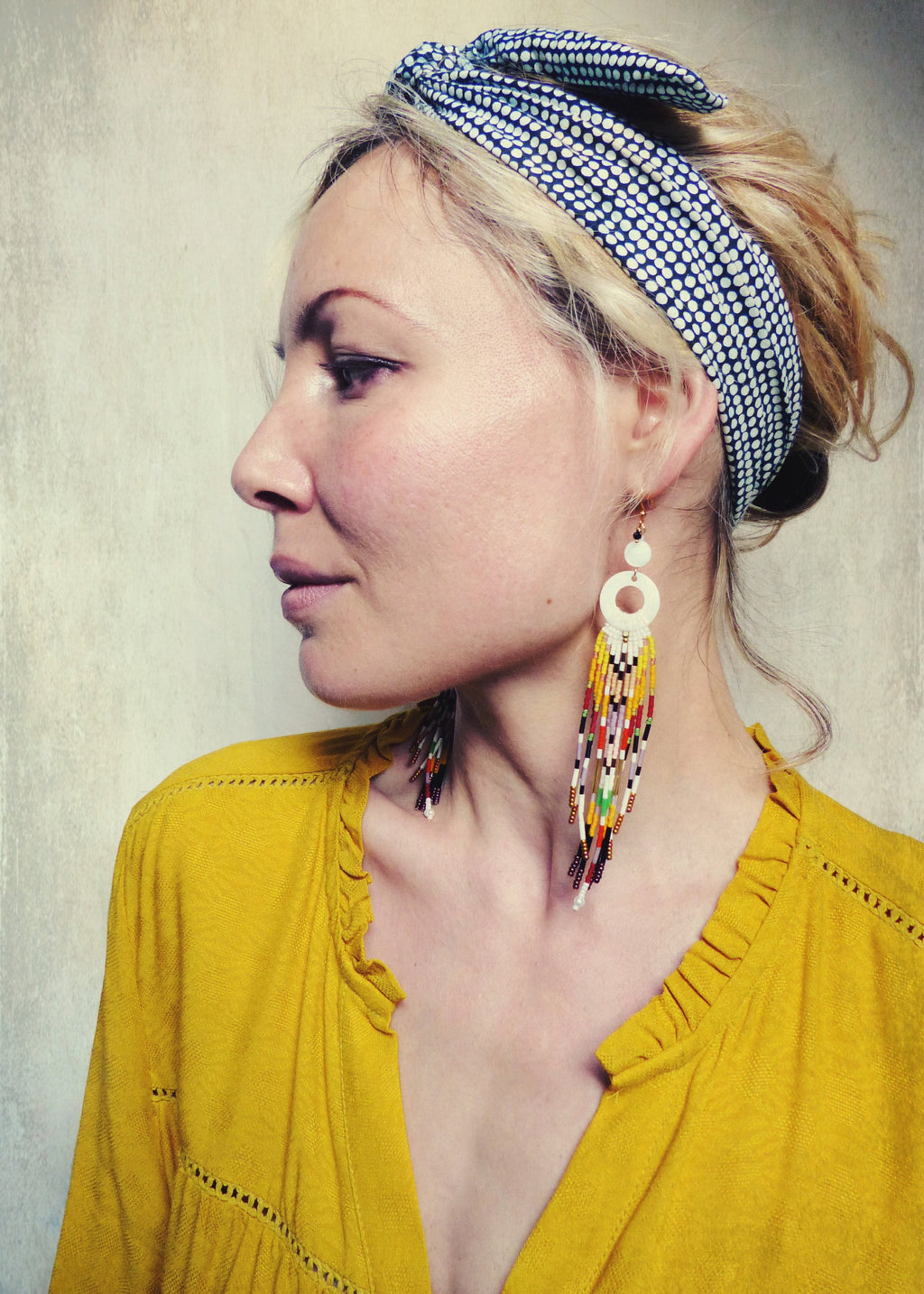 Peut inclure: Une femme portant un bandeau à pois bleu et blanc et un haut jaune à col en V profond. Elle porte de longues boucles d'oreilles en perles colorées avec un pendentif rond blanc.