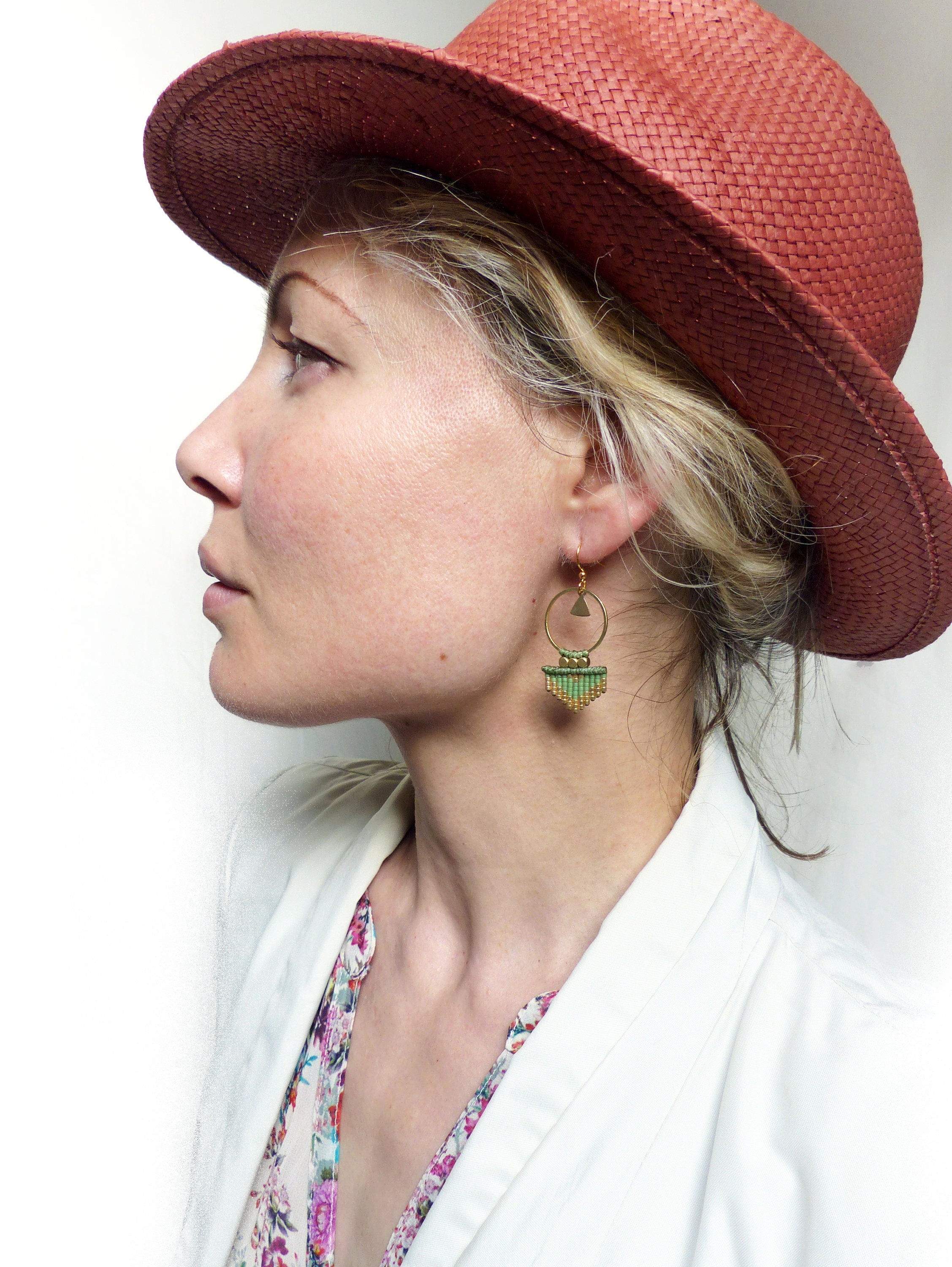 Peut inclure: Une femme portant un chapeau rouge et une veste blanche avec un chemisier à imprimé floral. Elle porte des boucles d'oreilles en perles vertes et dorées.