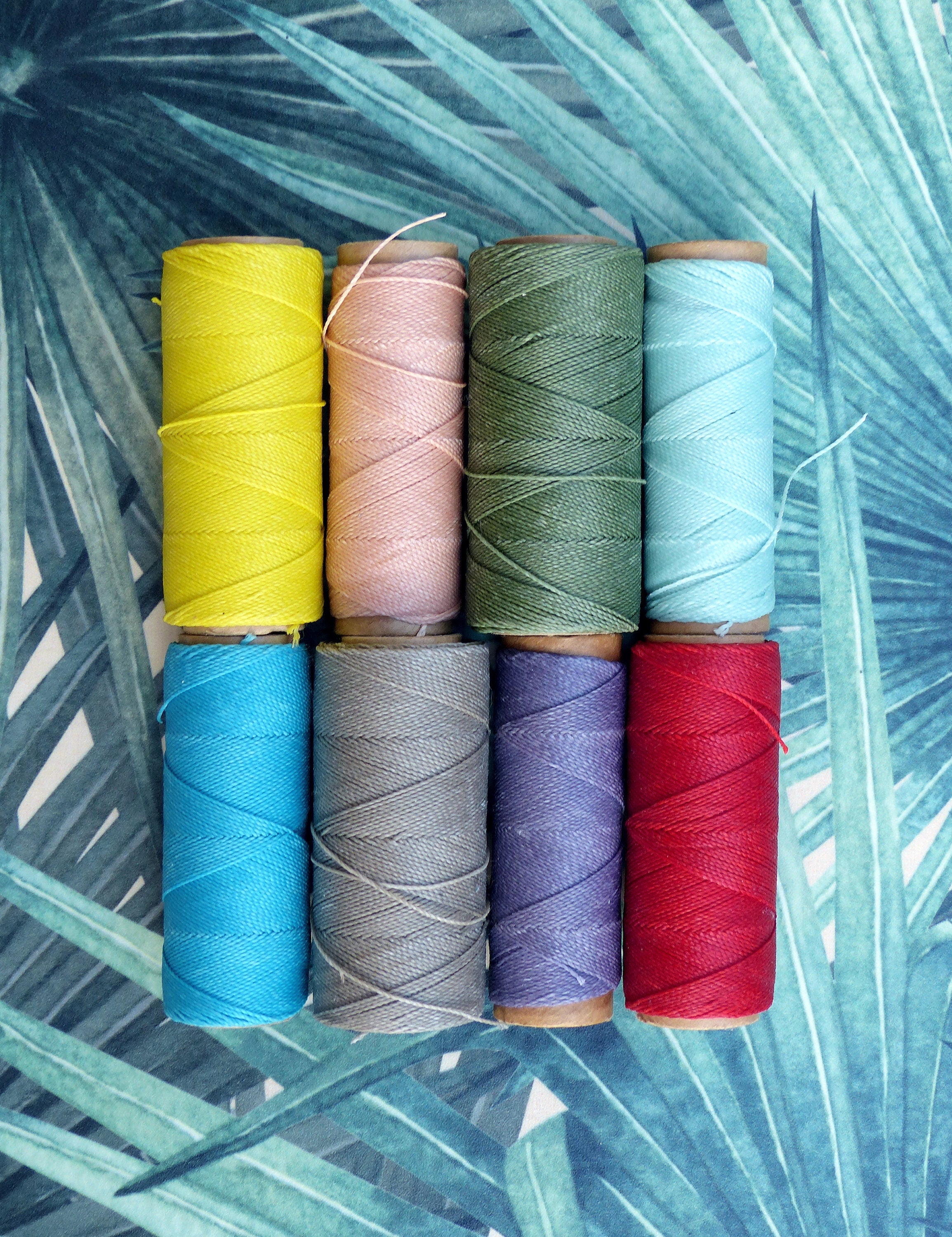 Peut inclure: Huit bobines de fil de différentes couleurs, dont jaune, rose, vert, bleu, gris, violet et rouge. Les bobines sont disposées en rangée sur un fond à motifs vert et blanc.
