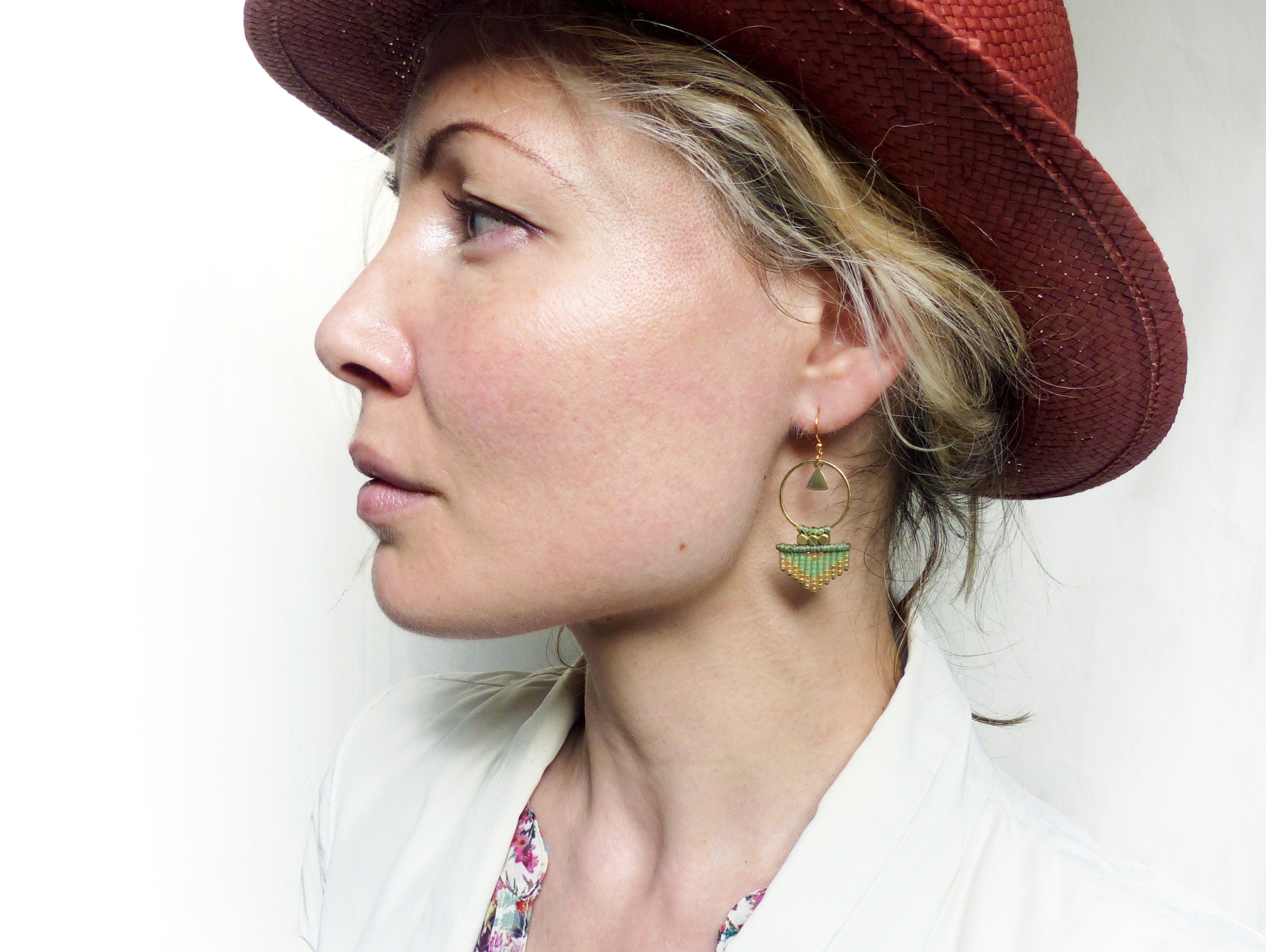Peut inclure: Une femme portant un chapeau à larges bords avec une paire de boucles d'oreilles pendantes géométriques vertes et dorées. Les boucles d'oreilles présentent un petit cercle avec un triangle à l'intérieur et une frange de perles vertes et dorées.
