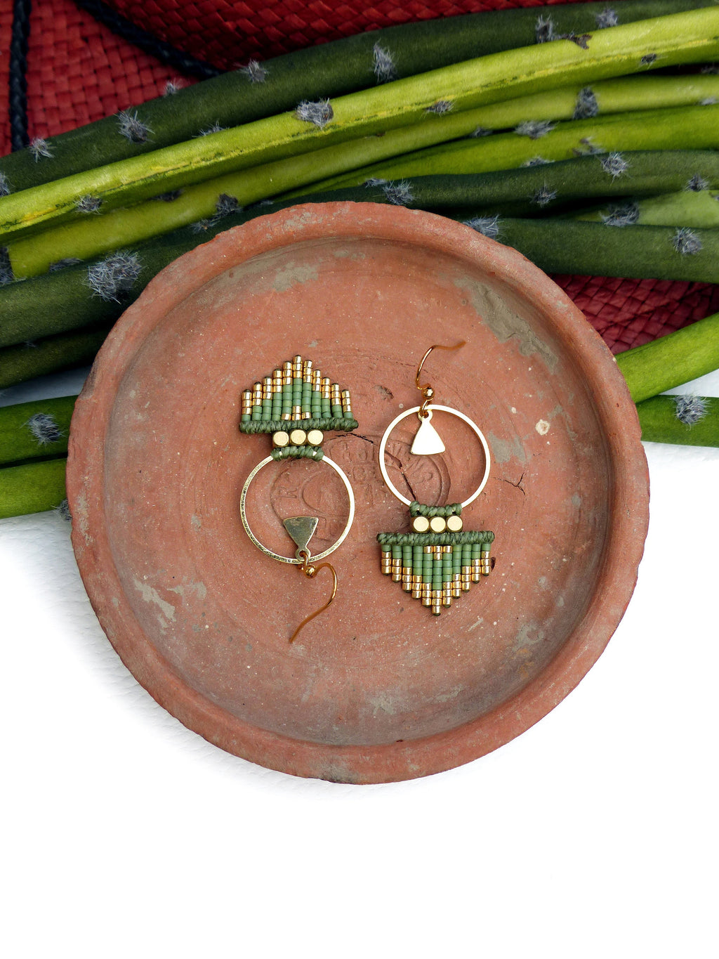 Peut inclure: Une paire de boucles d'oreilles en perles vertes et dorées avec un anneau et un pendentif triangulaire. Les boucles d'oreilles sont présentées sur une assiette en terre cuite.
