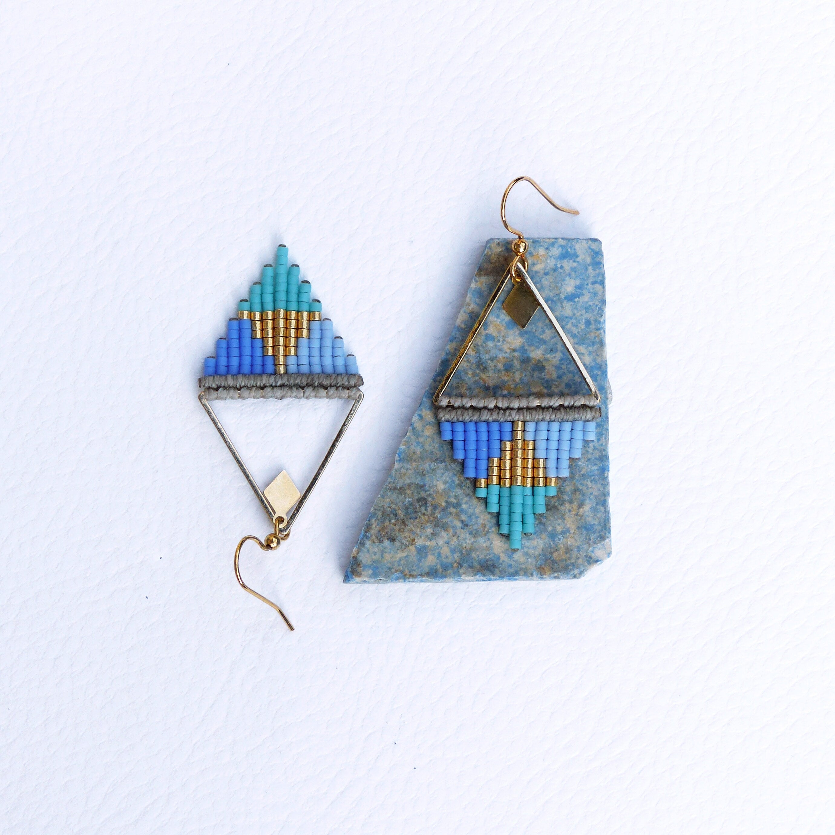 Peut inclure: Une paire de boucles d'oreilles triangulaires géométriques avec des perles bleues, turquoise et dorées. Les boucles d'oreilles sont suspendues à des crochets dorés.