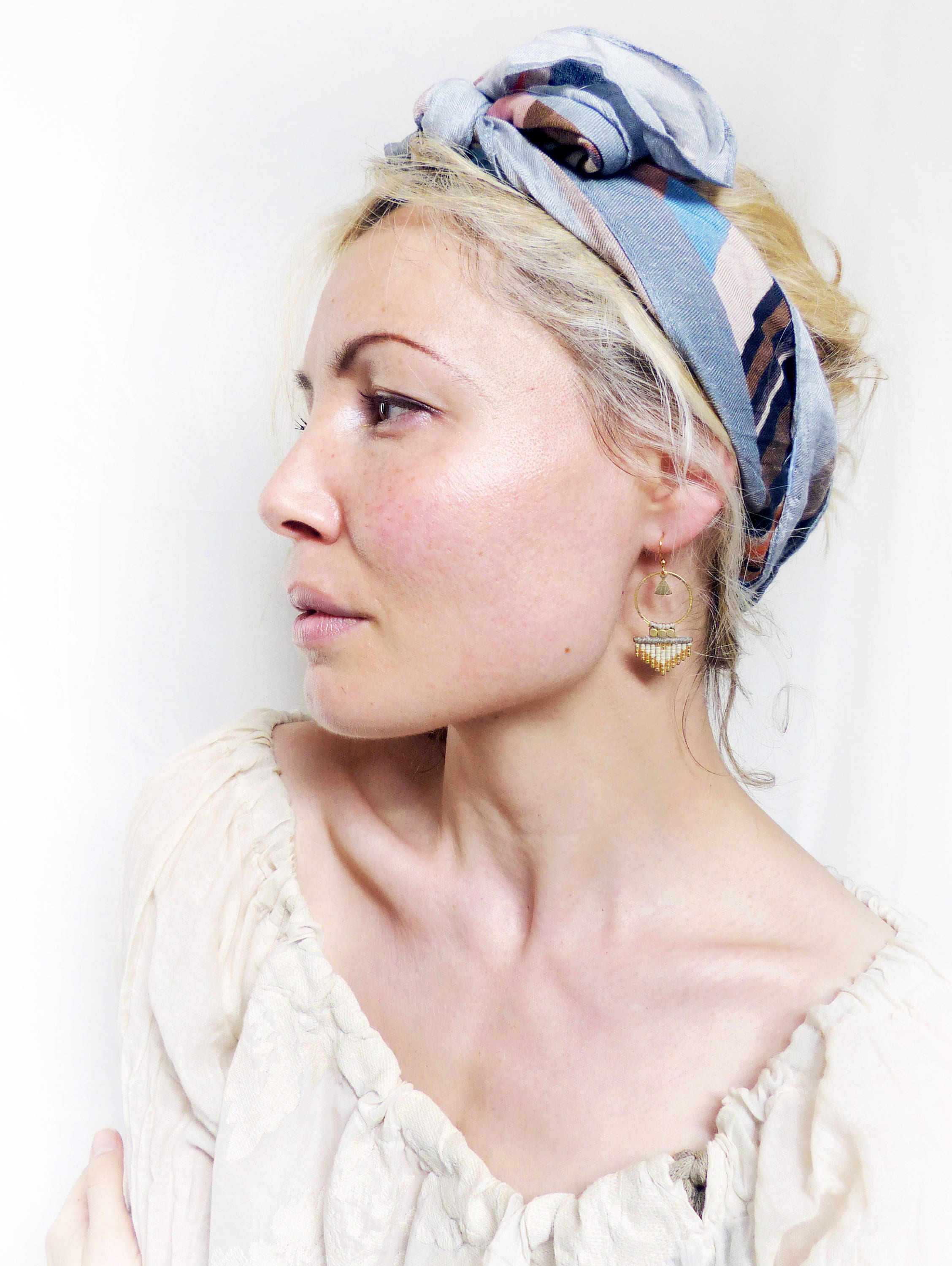 Peut inclure: Une femme aux cheveux blonds portant un foulard à motifs bleus et bruns et des boucles d'oreilles dorées. Elle porte un chemisier blanc à épaules dénudées.