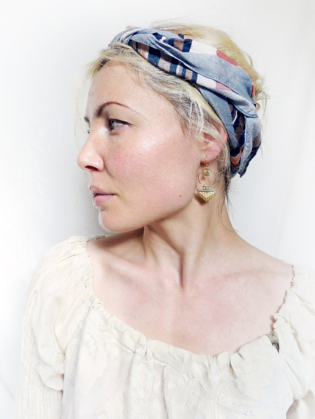 Peut inclure: Une femme aux cheveux blonds portant un foulard à motifs bleus et roses et des boucles d'oreilles créoles dorées avec un pendentif en perles. Elle porte une blouse blanche à col volanté.
