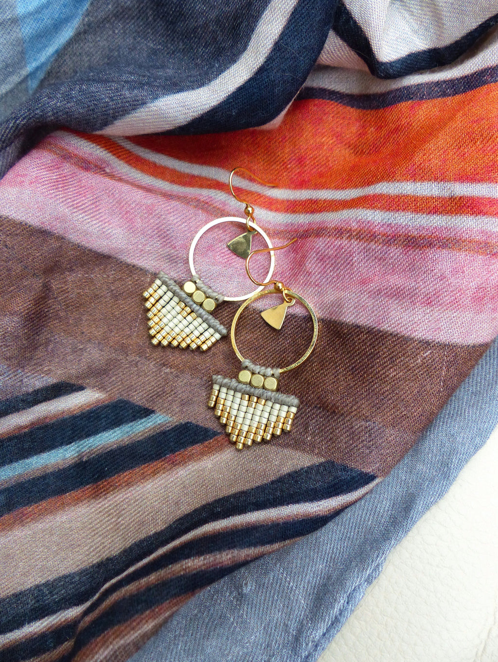 Peut inclure: Une paire de boucles d'oreilles créoles dorées avec une frange de perles. La frange est faite de perles blanches et dorées et a la forme d'une larme. Les boucles d'oreilles sont suspendues à un fond de tissu rayé.