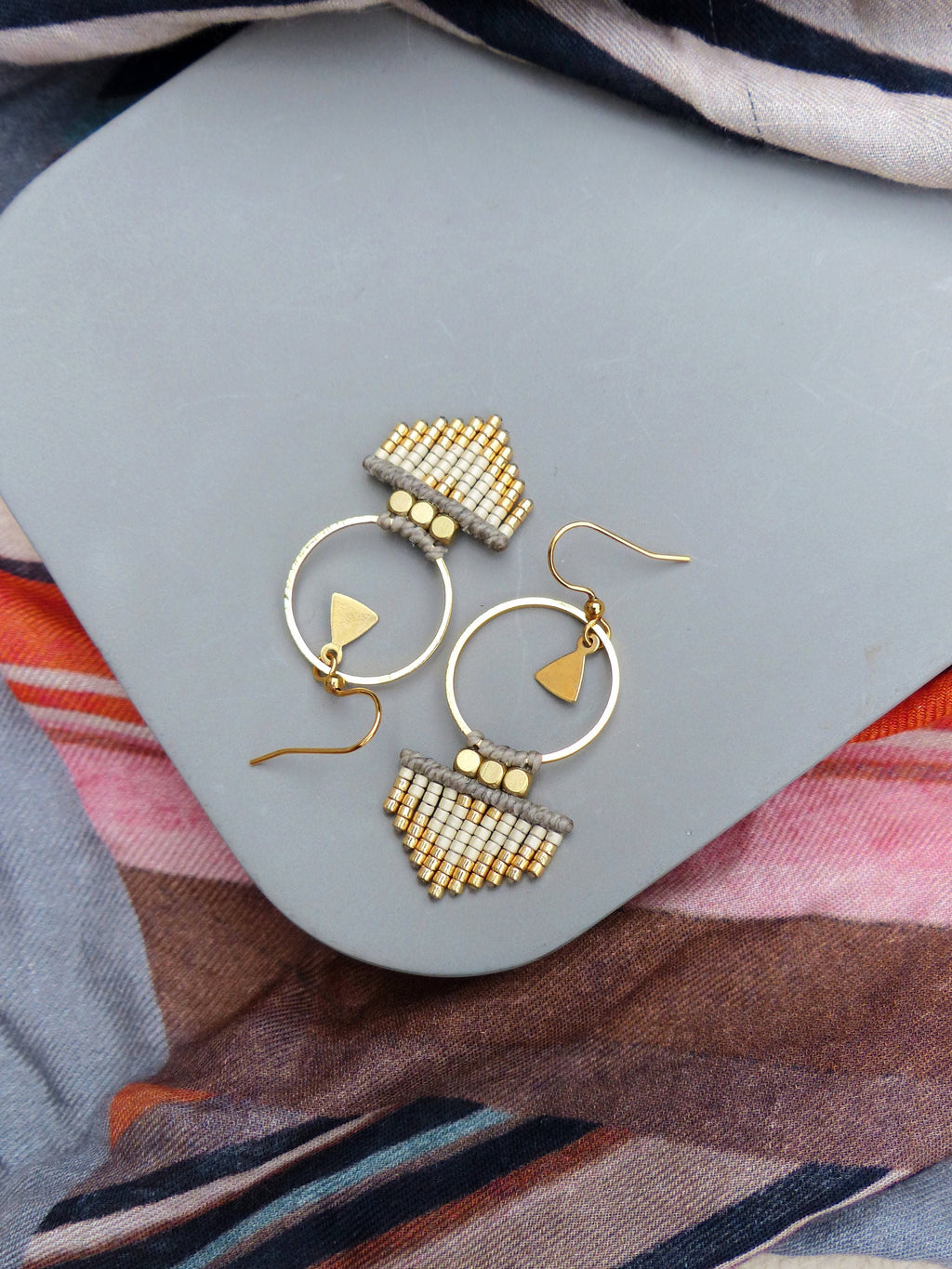 Peut inclure: Une paire de boucles d'oreilles créoles dorées avec une frange de perles blanches et dorées. Les boucles d'oreilles sont sur une surface grise.