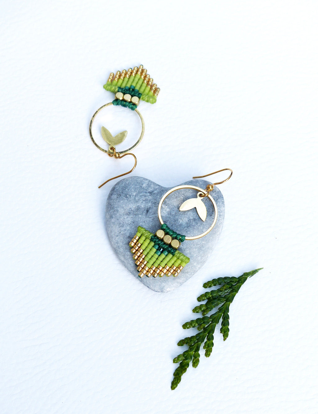 Peut inclure: Une paire de boucles d'oreilles en perles vertes et dorées avec un anneau en or et un charme en forme de feuille. Les boucles d'oreilles sont présentées sur une pierre grise.