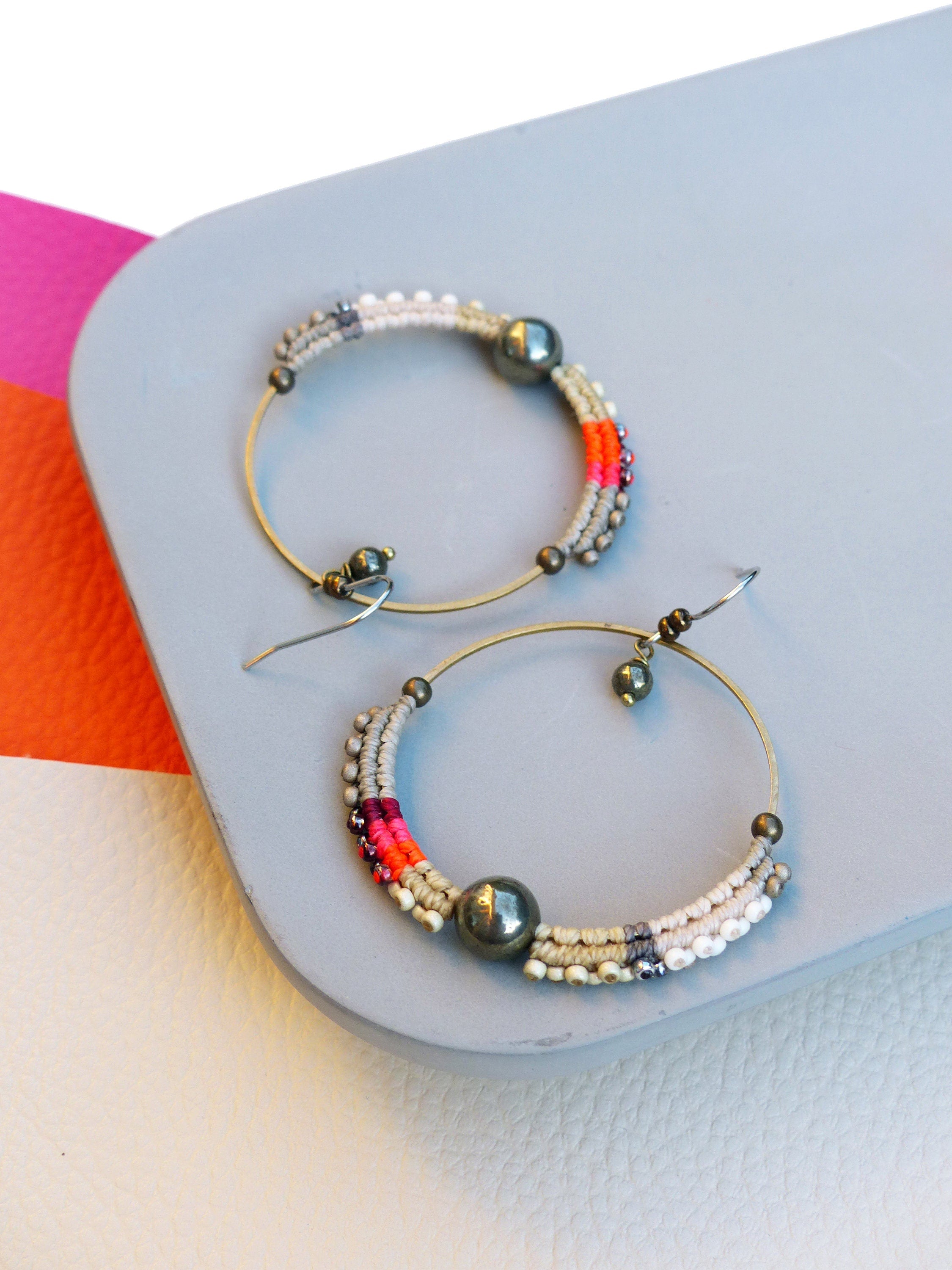 Peut inclure: Deux boucles d'oreilles créoles dorées. Chaque boucle d'oreille présente une section de fil tissé multicolore en blanc, gris, rose et orange, avec une grosse perle gris foncé. Les boucles d'oreilles sont posées sur une surface grise.