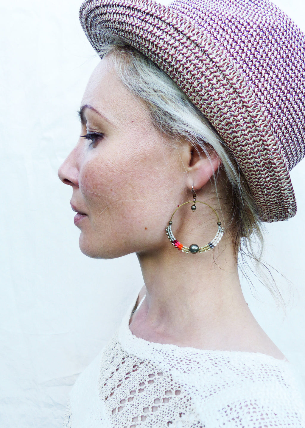 Peut inclure: Une femme porte un chapeau de paille avec un motif tissé marron et blanc et de grandes boucles d'oreilles créoles. Les boucles d'oreilles sont dorées et ornées de perles blanches, rouges et noires. La femme porte un haut blanc en maille ajourée.